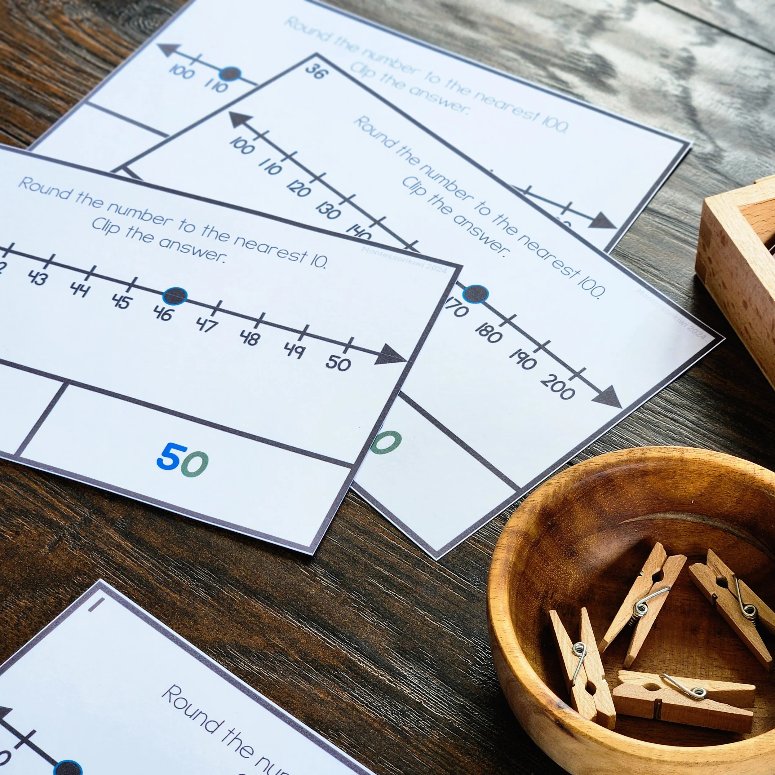 Close-up of a worksheet with arithmetic problems involving rounding numbers to the nearest 10, 100, and 1000 on a wooden surface, with a wooden bowl containing clothespins and some wooden clothespin holders nearby.
