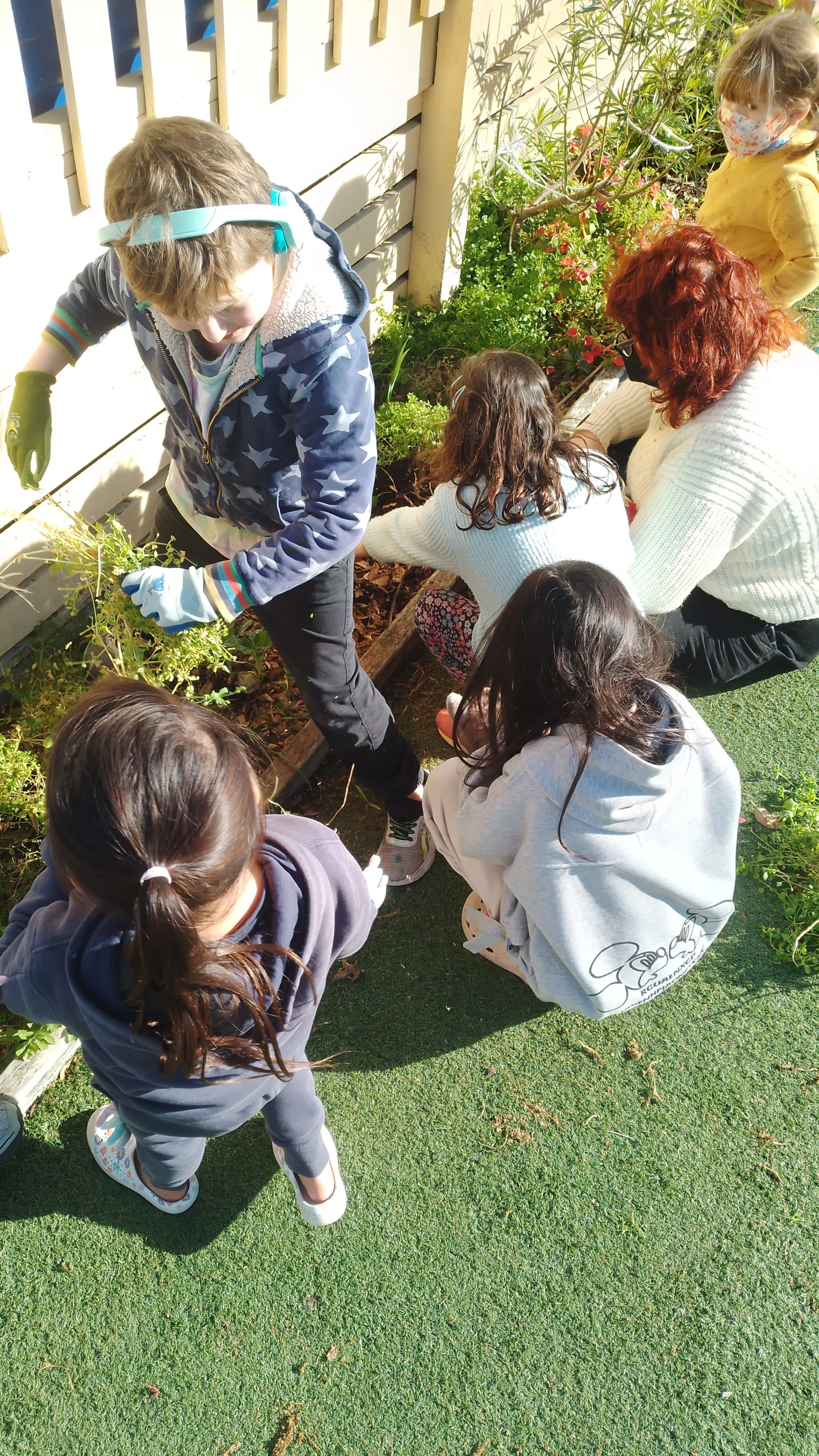 Children and an adult gardening together outdoors, planting or weeding in a garden bed beside a wooden fence, some children wearing masks.