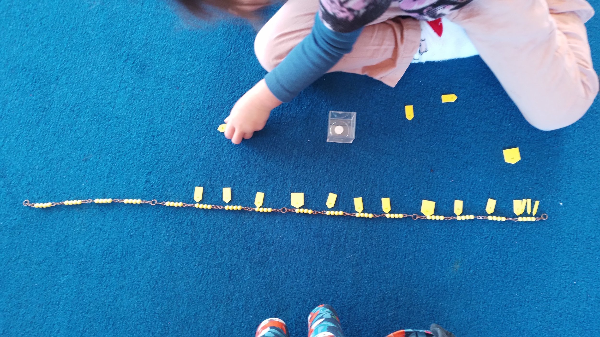 A child sitting on a blue carpet, playing with yellow puzzle pieces and a string of beads with yellow beads. The child's hand is reaching toward the puzzle pieces. Learning multiplication basic facts.