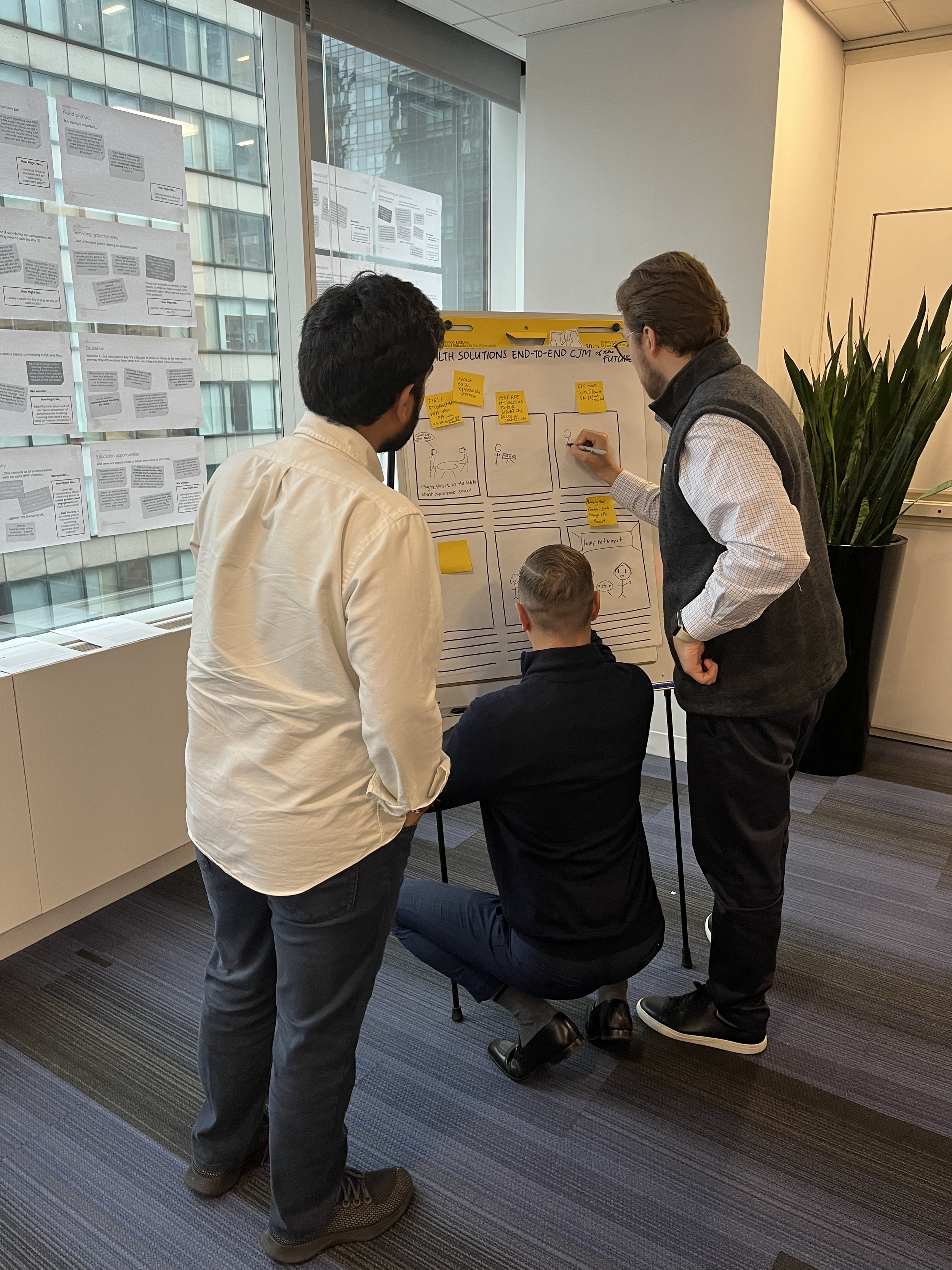 Three men working on a project at a whiteboard in a modern office with large windows and cityscape view. One man is sitting, while the other two are standing and discussing.