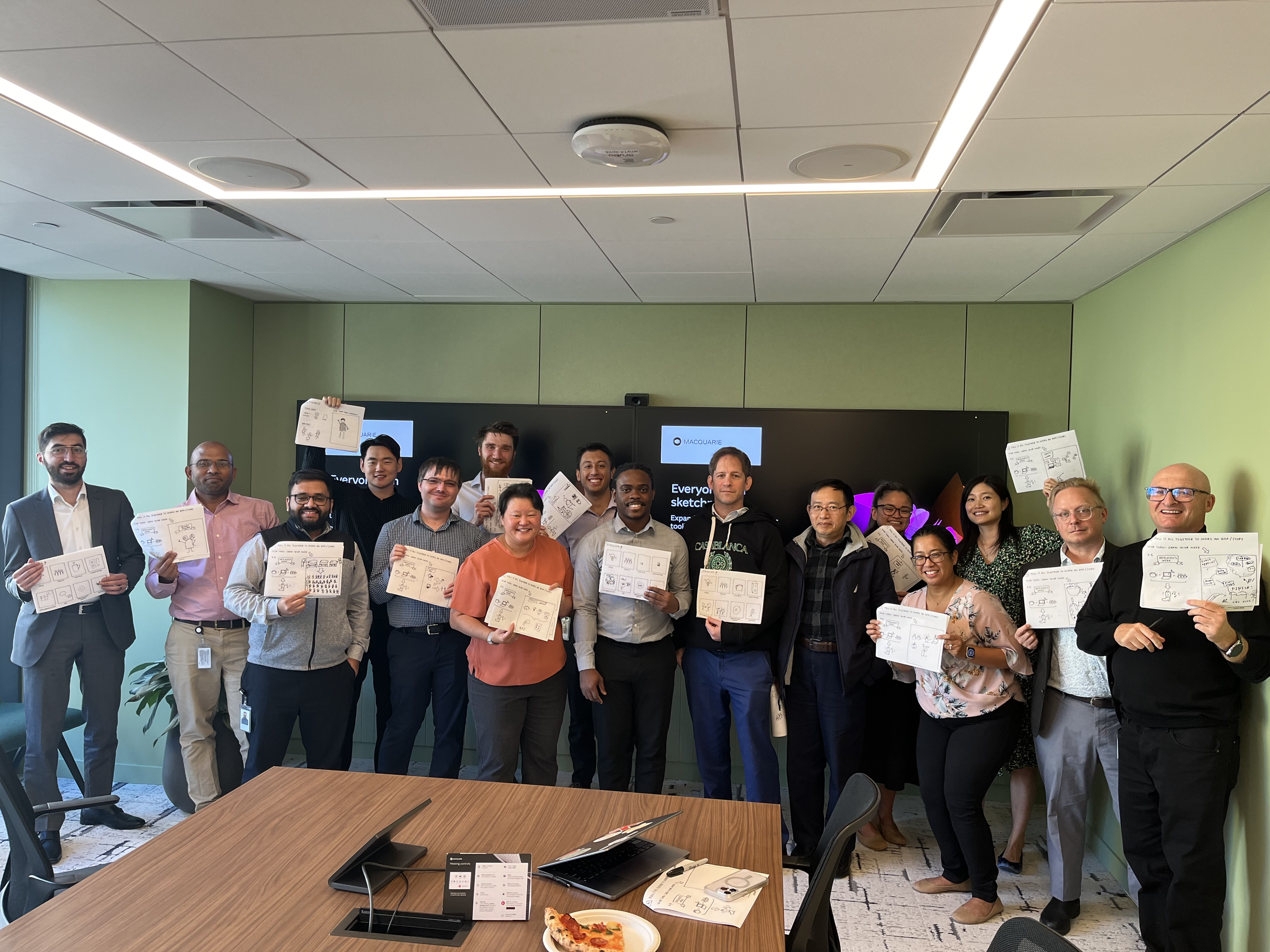 Group of diverse professionals in a conference room holding sketch papers with drawings, standing in front of a screen with a presentation.