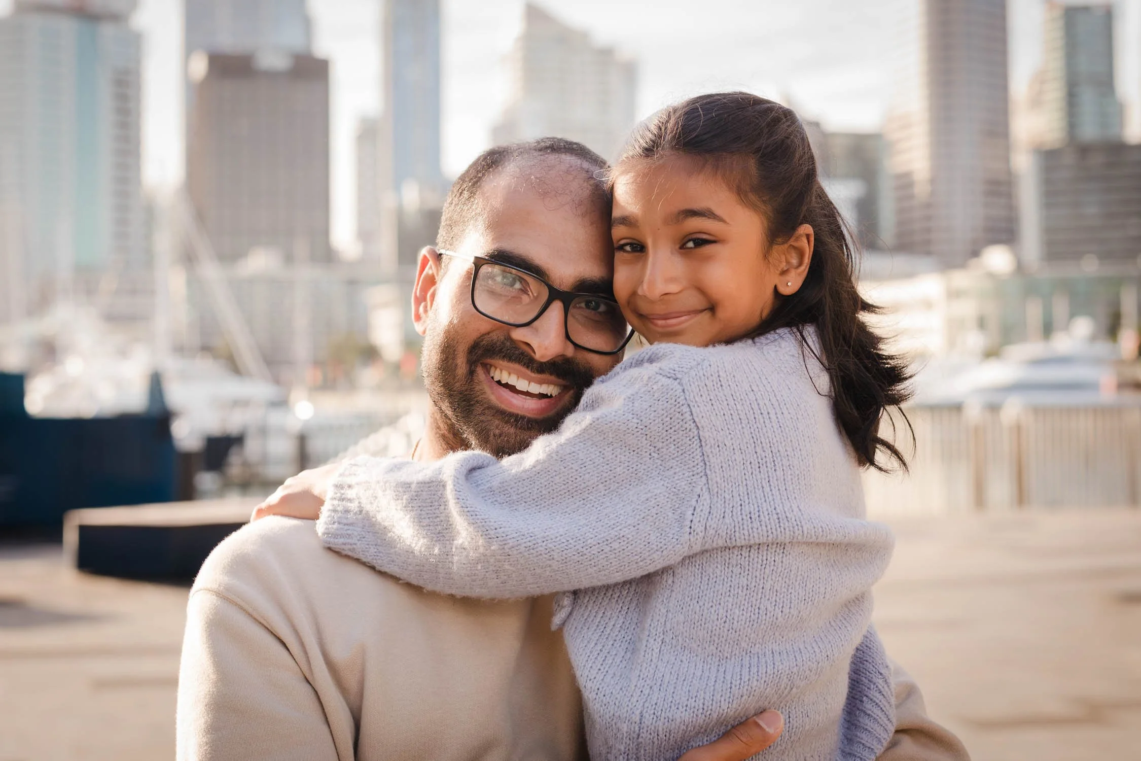 A man with glasses and a beard holding a young girl in a sweater, both smiling happily, with city buildings and a marina in the background.