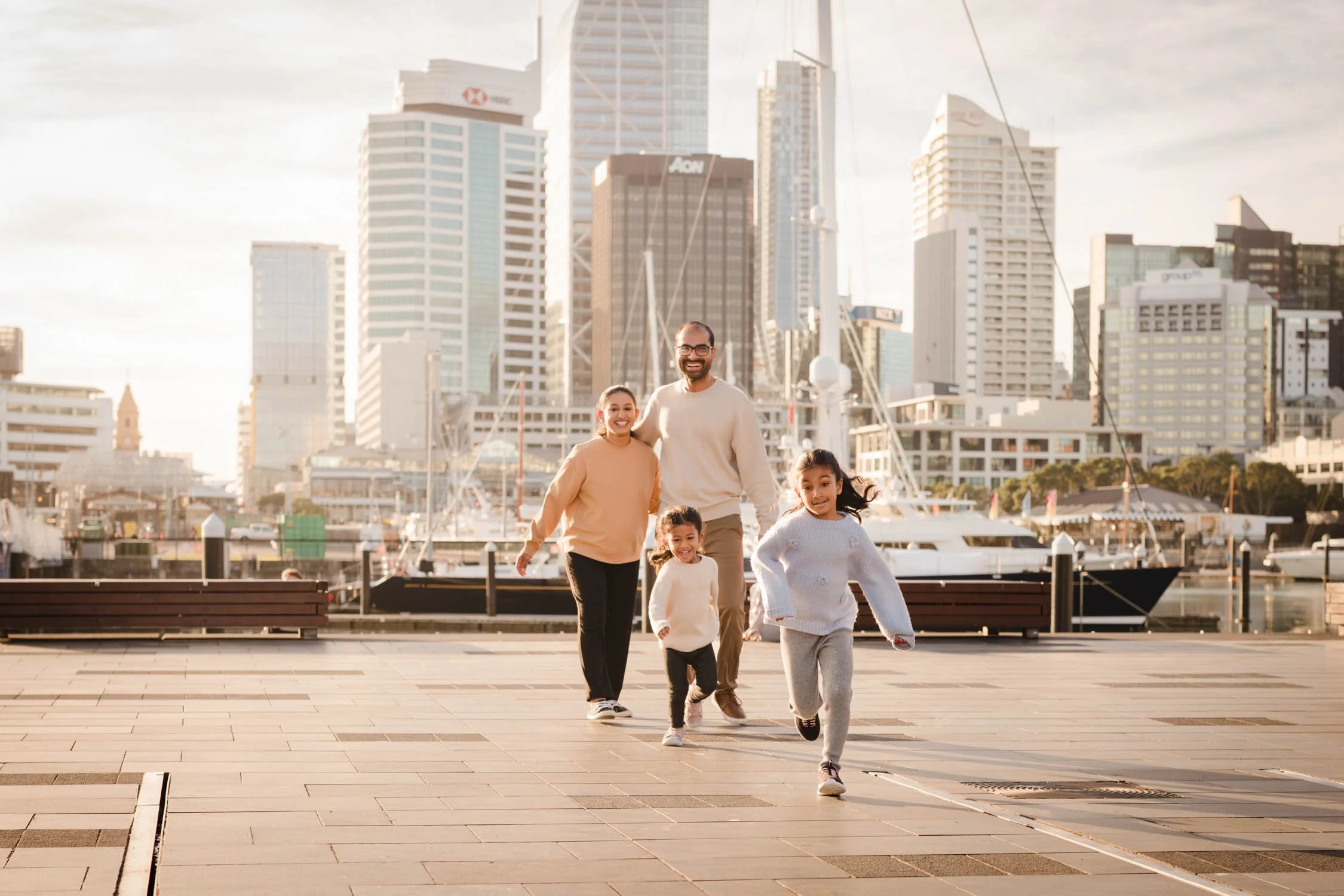 A family of five, with adults and children, running along a waterfront promenade with boats in the harbor and a skyline of tall modern buildings in the background, during sunset.
