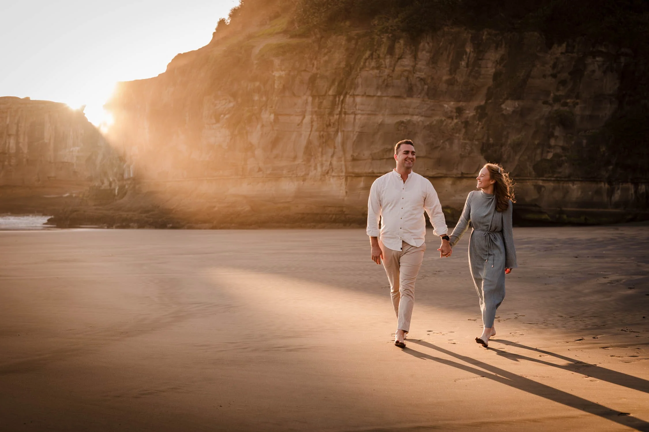 A smiling man and woman holding hands while walking on a sandy beach at sunset, with cliffs in the background.