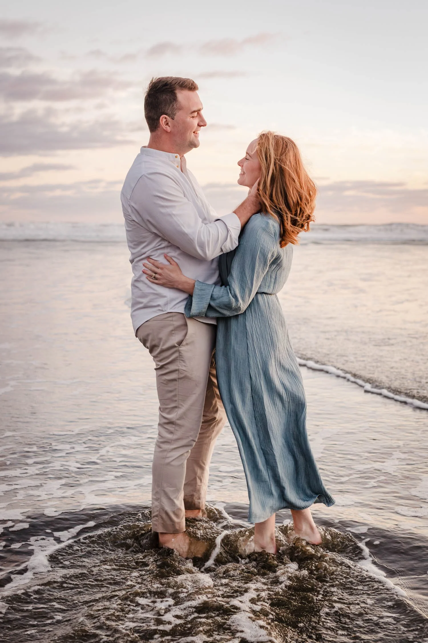 A couple standing in the water at the beach, smiling and looking at each other during sunset.