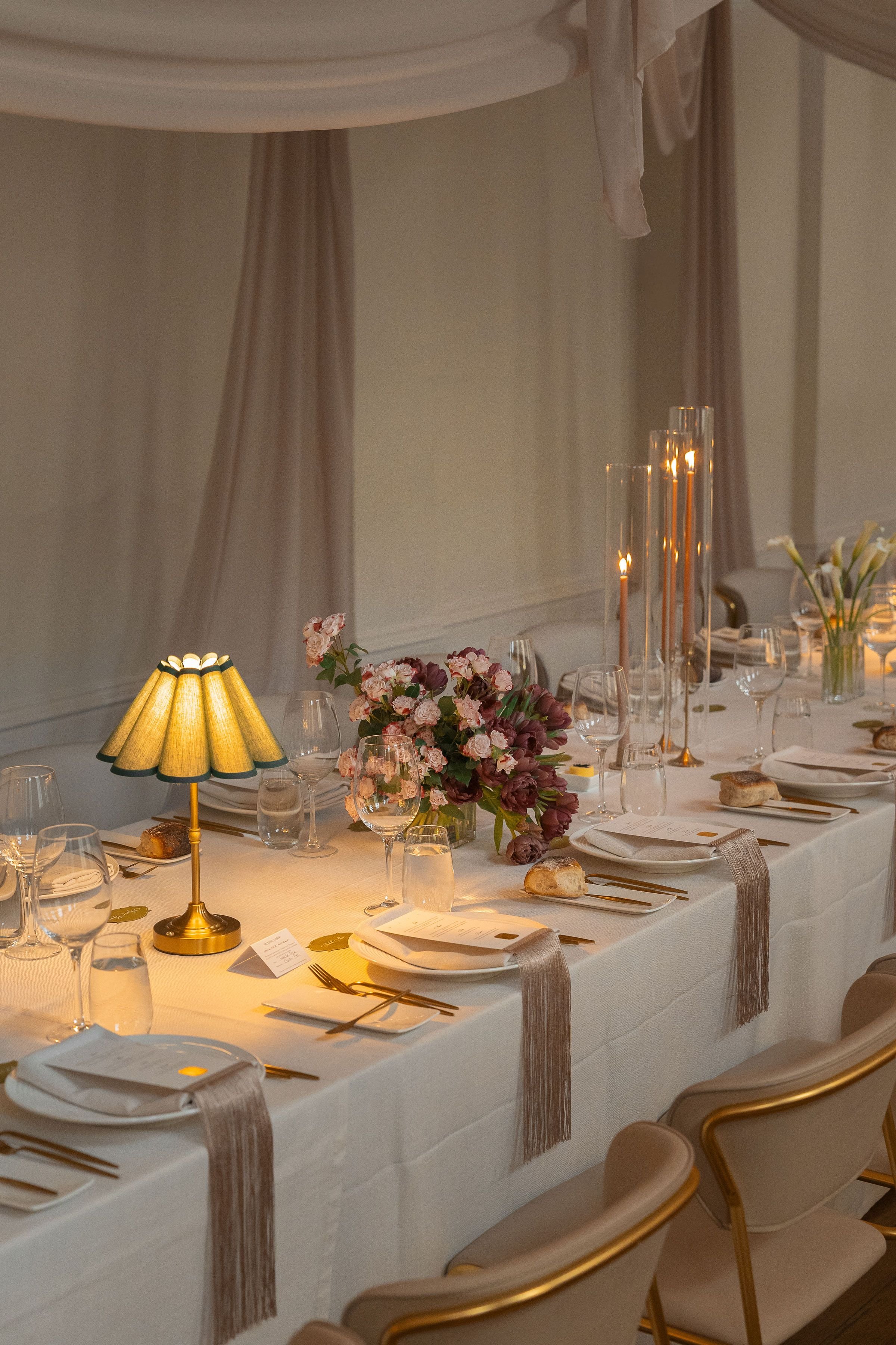 Elegant dining table set for a formal event, decorated with floral arrangements, candles, and fine tableware, with soft lighting and drapes in the background.
