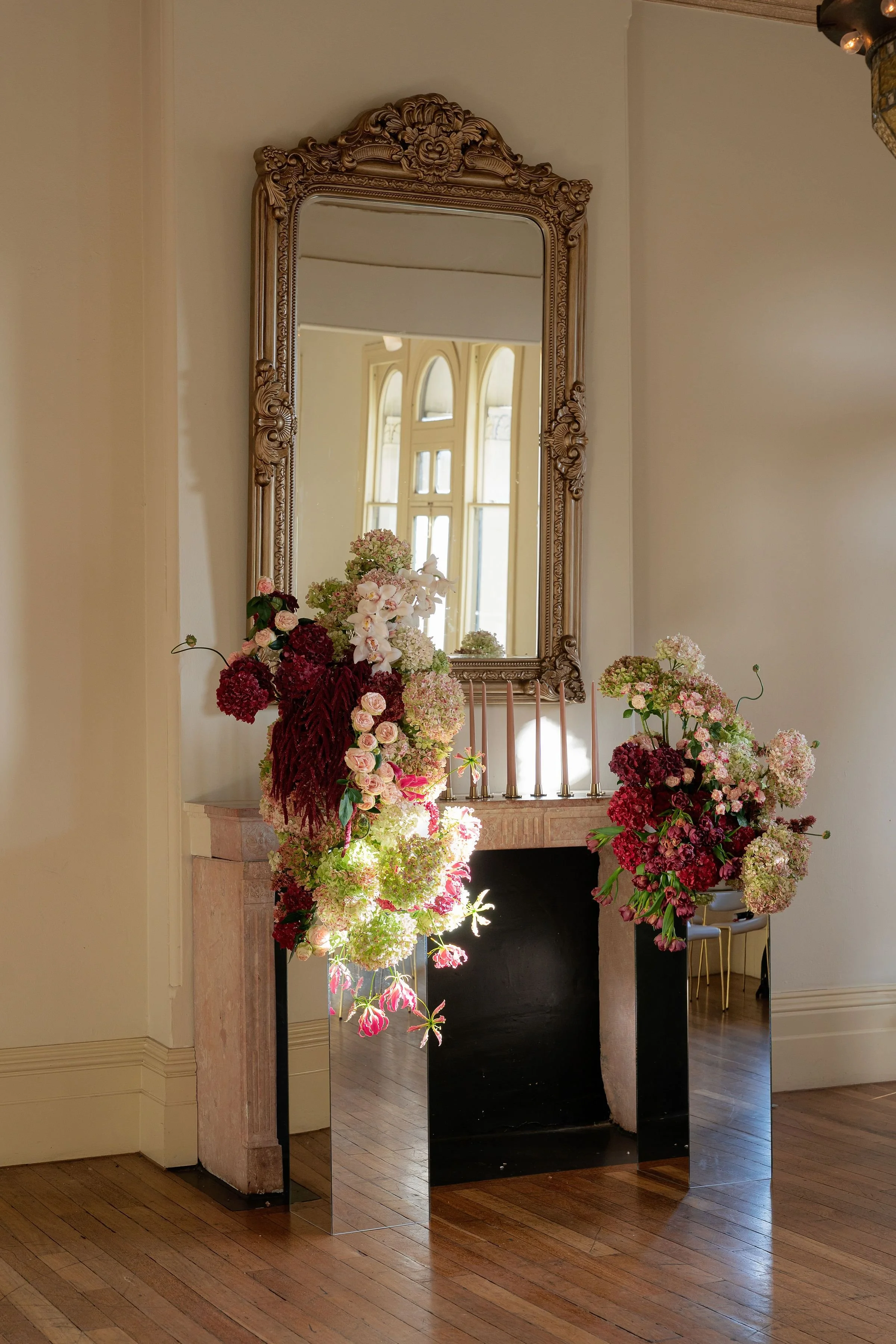 An ornate mirror hanging above a fireplace, with flower arrangements on either side, situated in a room with hardwood floors and tall windows in the background.
