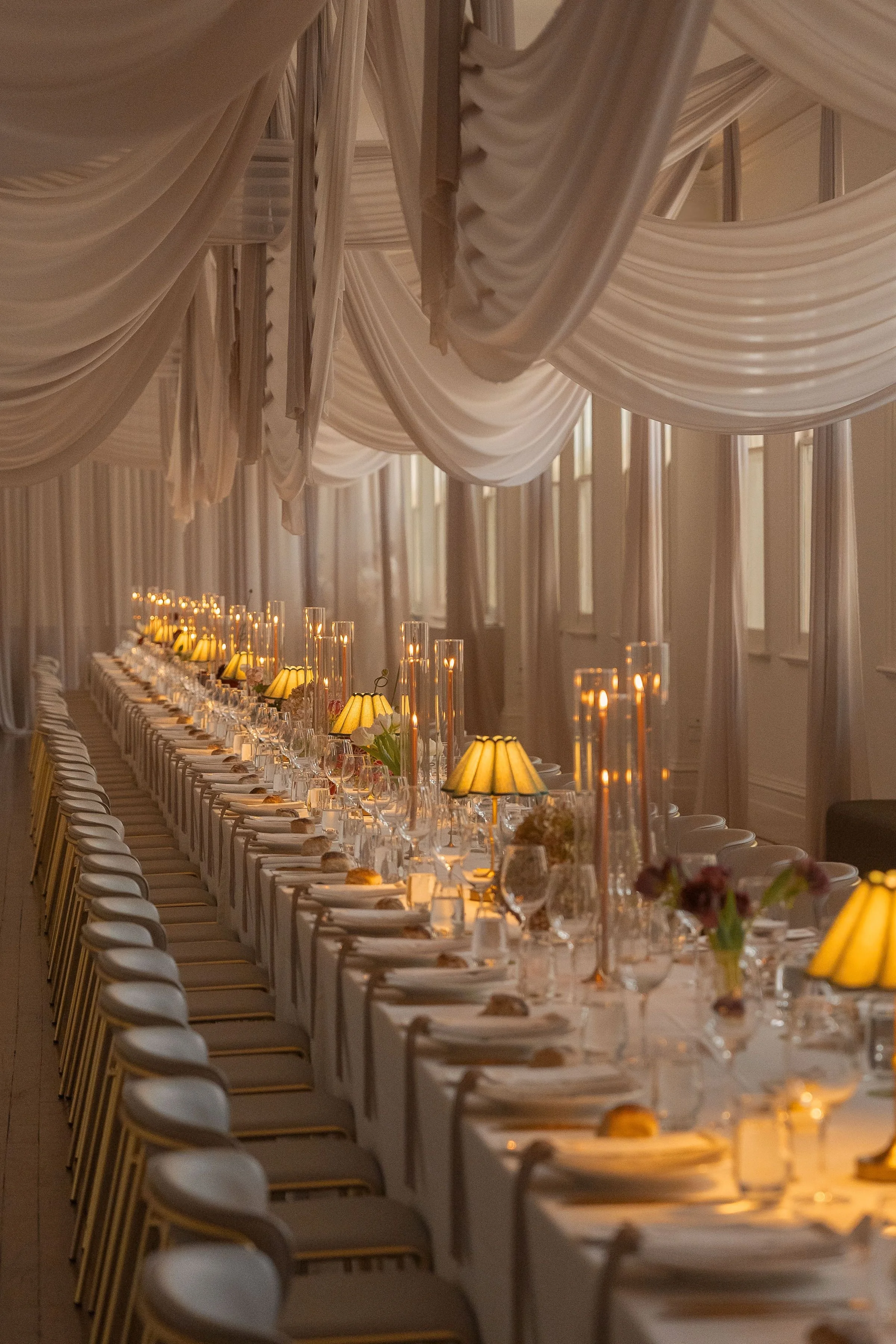 A long banquet table set for a formal event with white tablecloths, folded napkins, flowers, and yellow lampshades with candles inside. The room has draped beige fabric hanging from the ceiling.