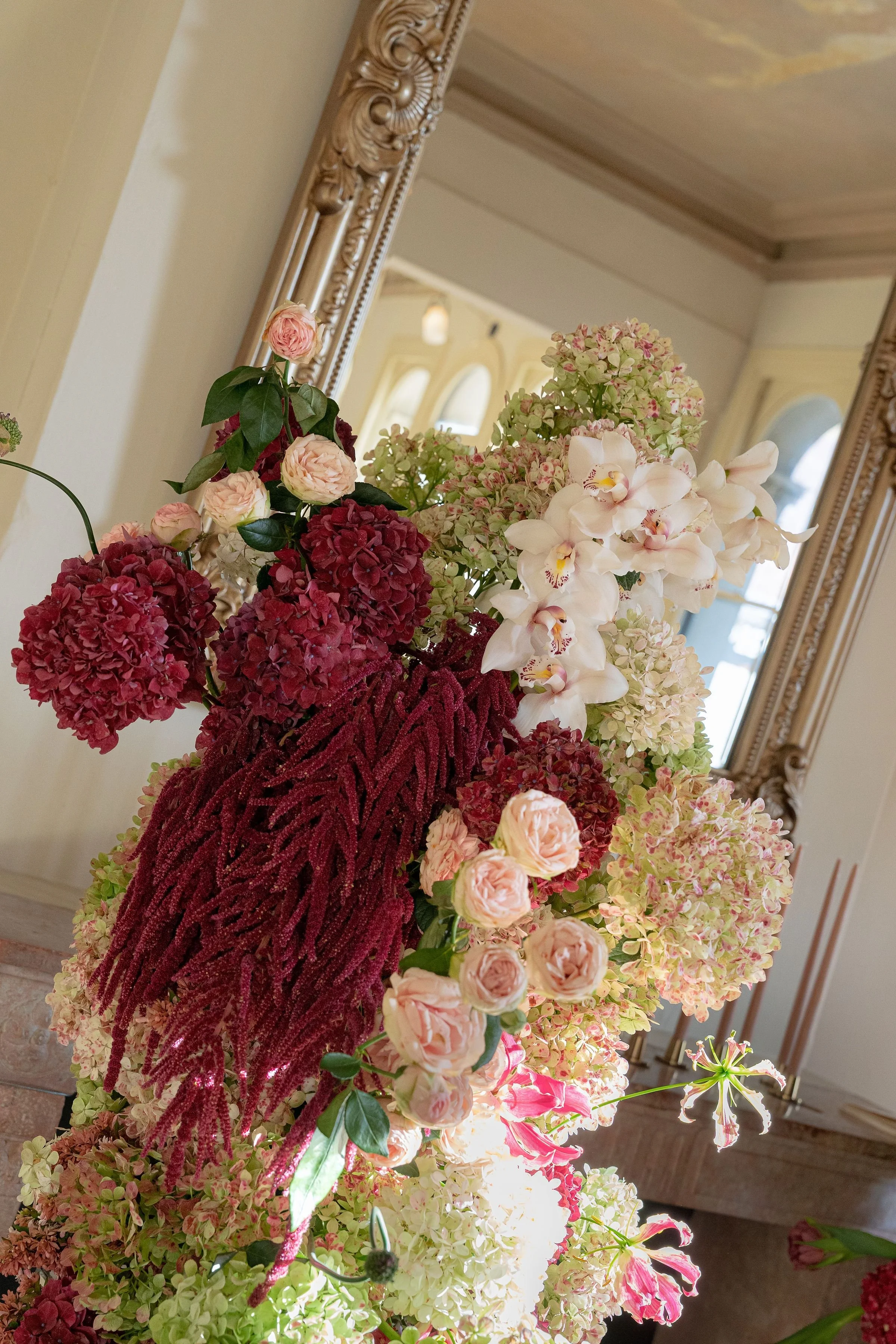 A colorful arrangement of flowers including pink roses, red hydrangeas, white orchids, and pink hydrangeas, placed in front of an ornate mirror in a bright room.