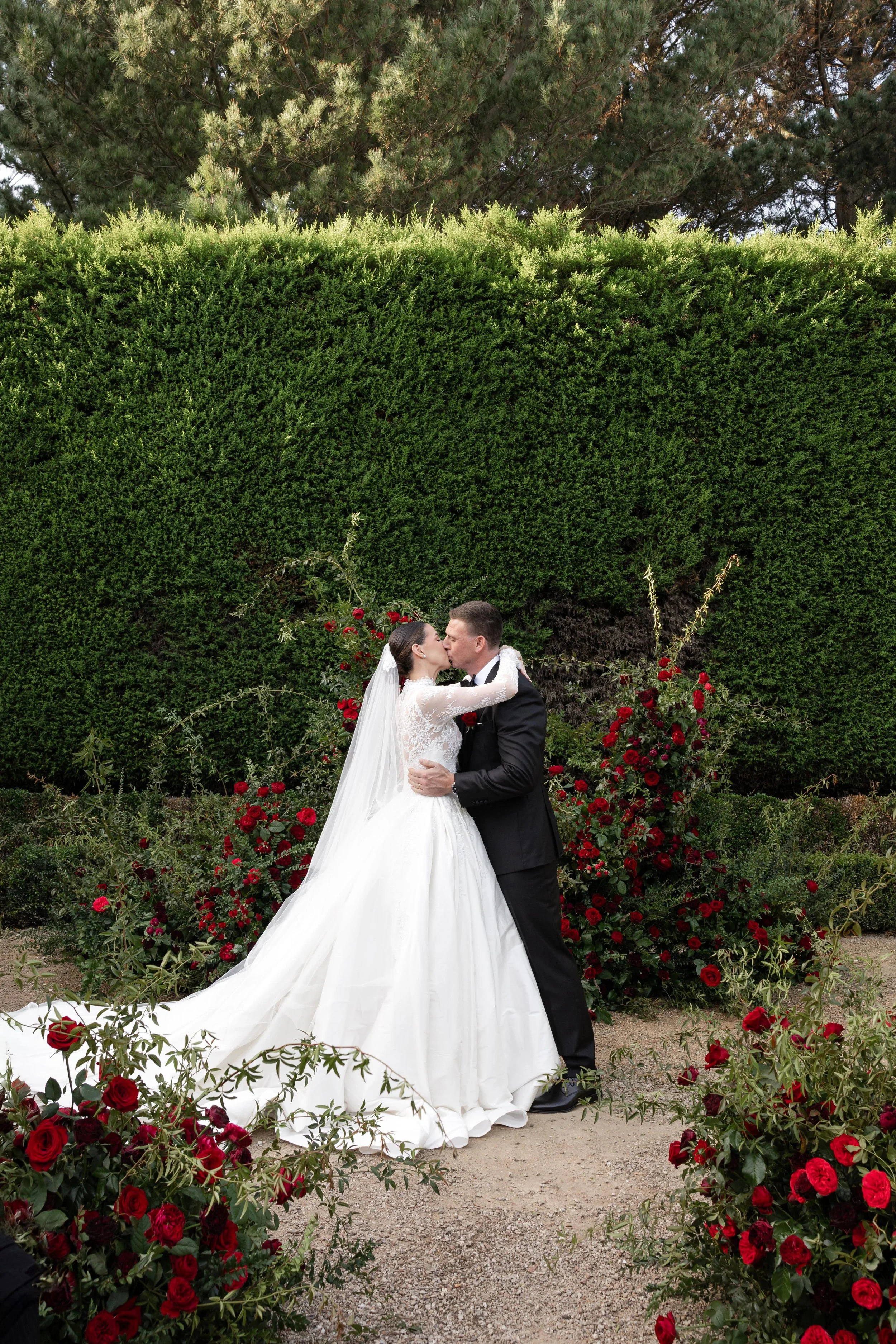 A bride and groom kiss outdoors amid red roses and green bushes with tall trees in the background.