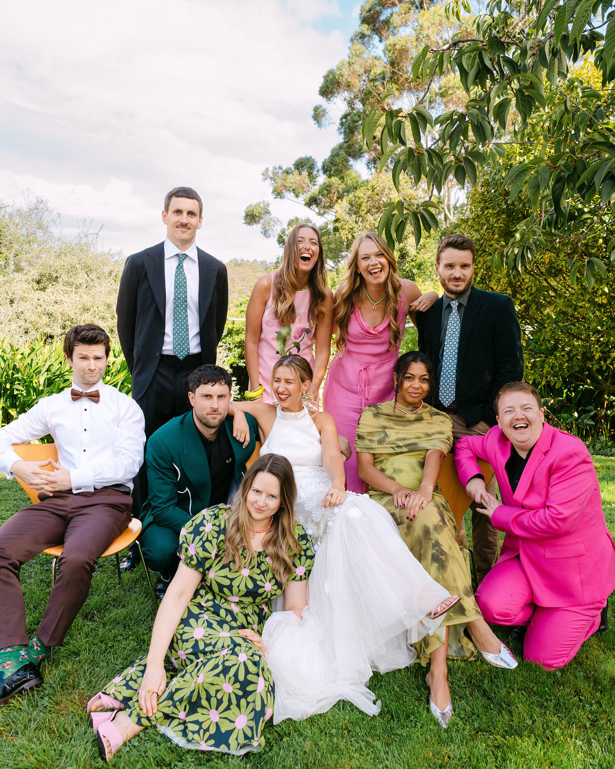 A lively wedding party poses in front of a grand, weathered turquoise door with "Temperance Hall" engraved above it. The bridesmaids, dressed in soft lilac and pastel shades, hold colorful bouquets and strike playful poses. 