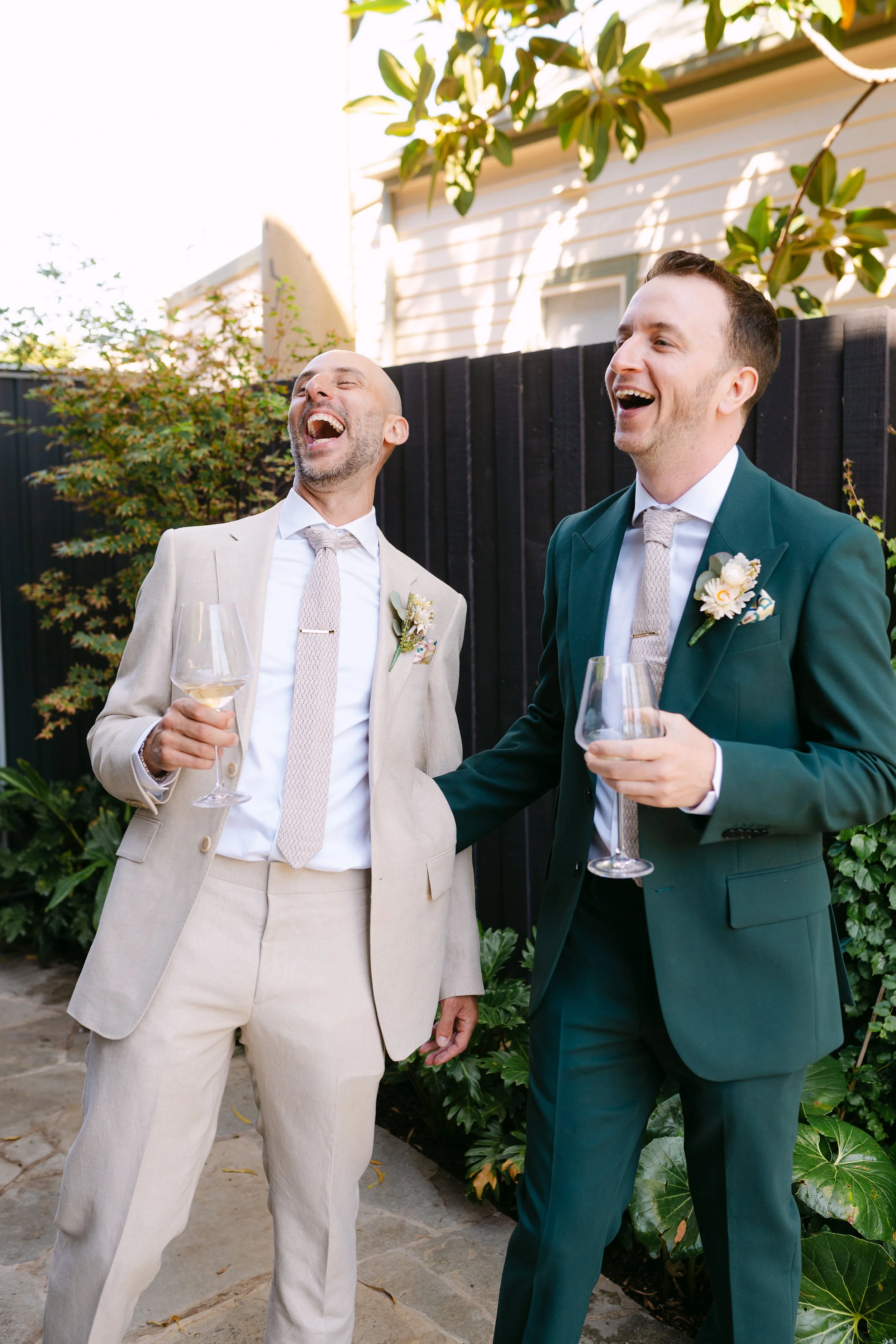  A bride in a white off-shoulder dress with her back to the camera, revealing a detailed tattoo on her upper back, faces her father crying at her in the wedding dress wearing a beige suit.