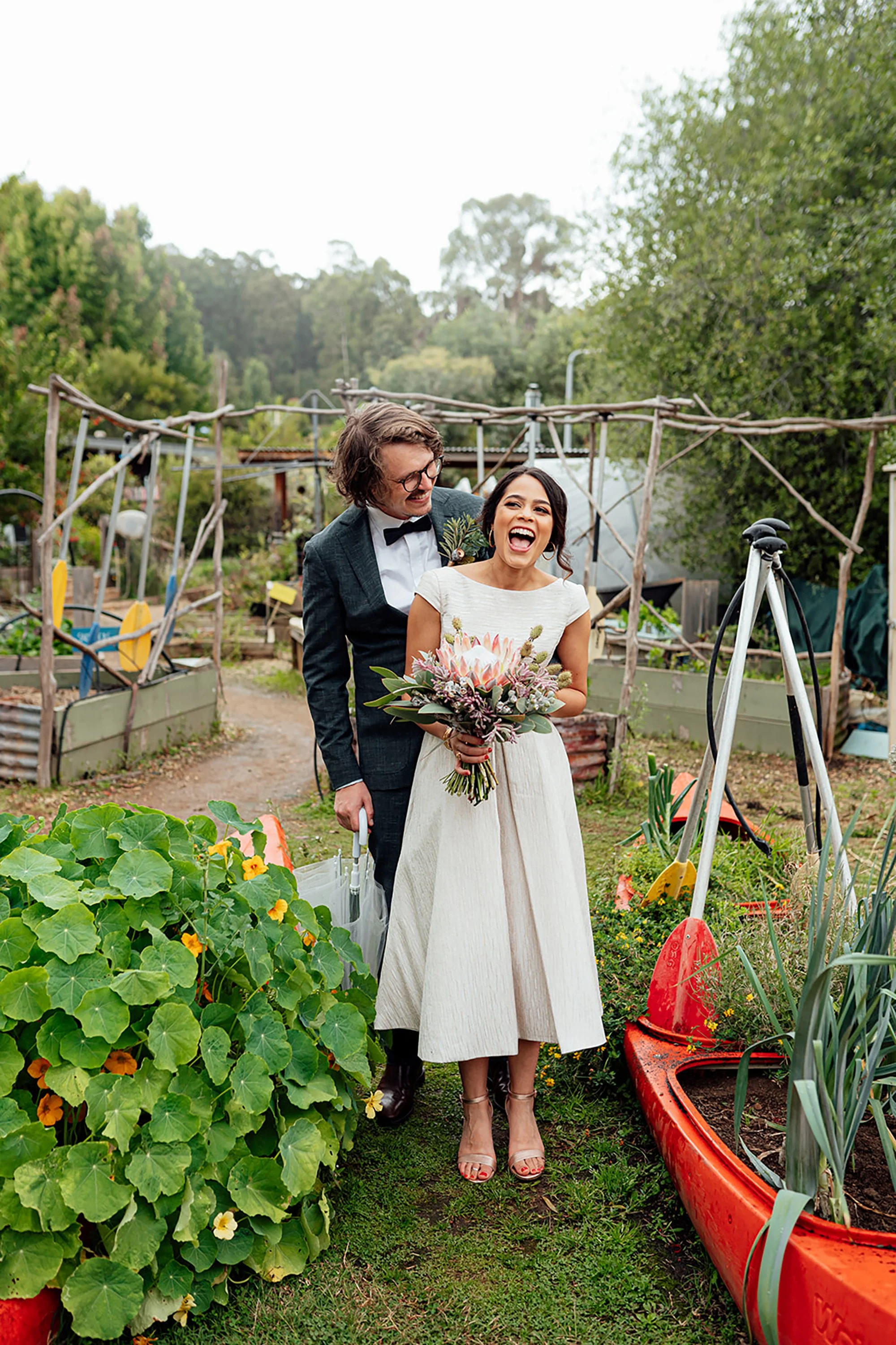 A newlywed couple walking through a rustic garden, with the groom in a black suit and the bride holding a bouquet, both smiling.