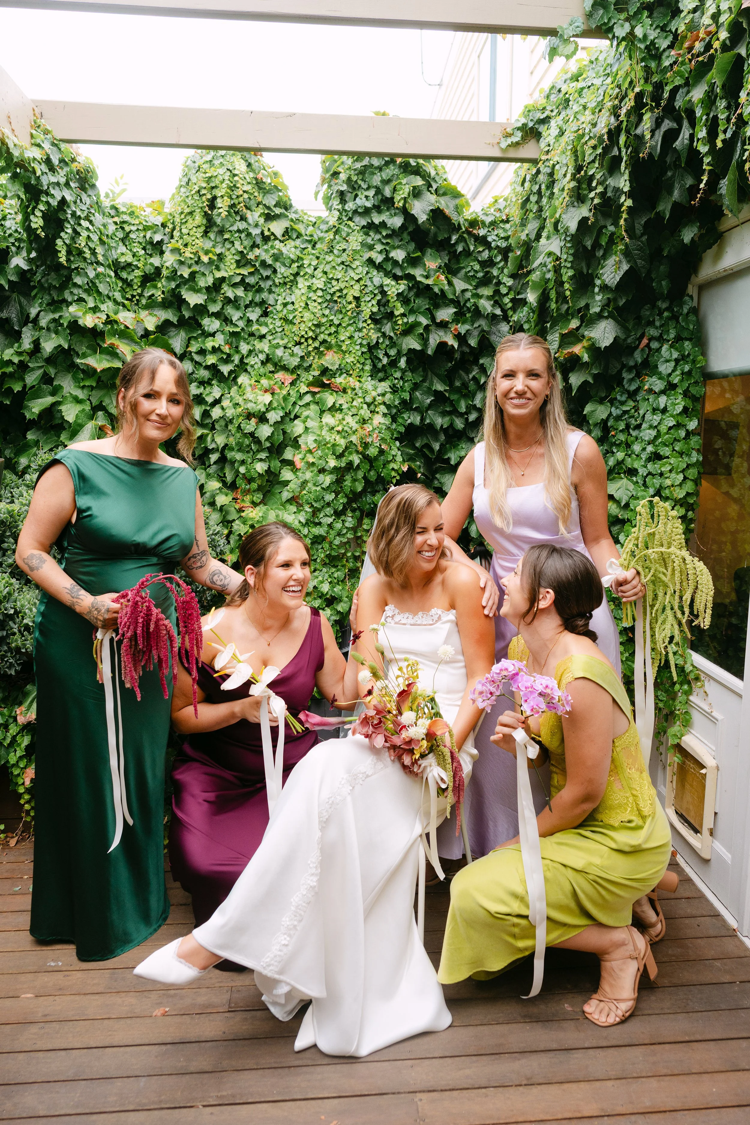 A lively wedding party poses in front of a grand, weathered turquoise door with "Temperance Hall" engraved above it. The bridesmaids, dressed in soft lilac and pastel shades, hold colorful bouquets and strike playful poses. 