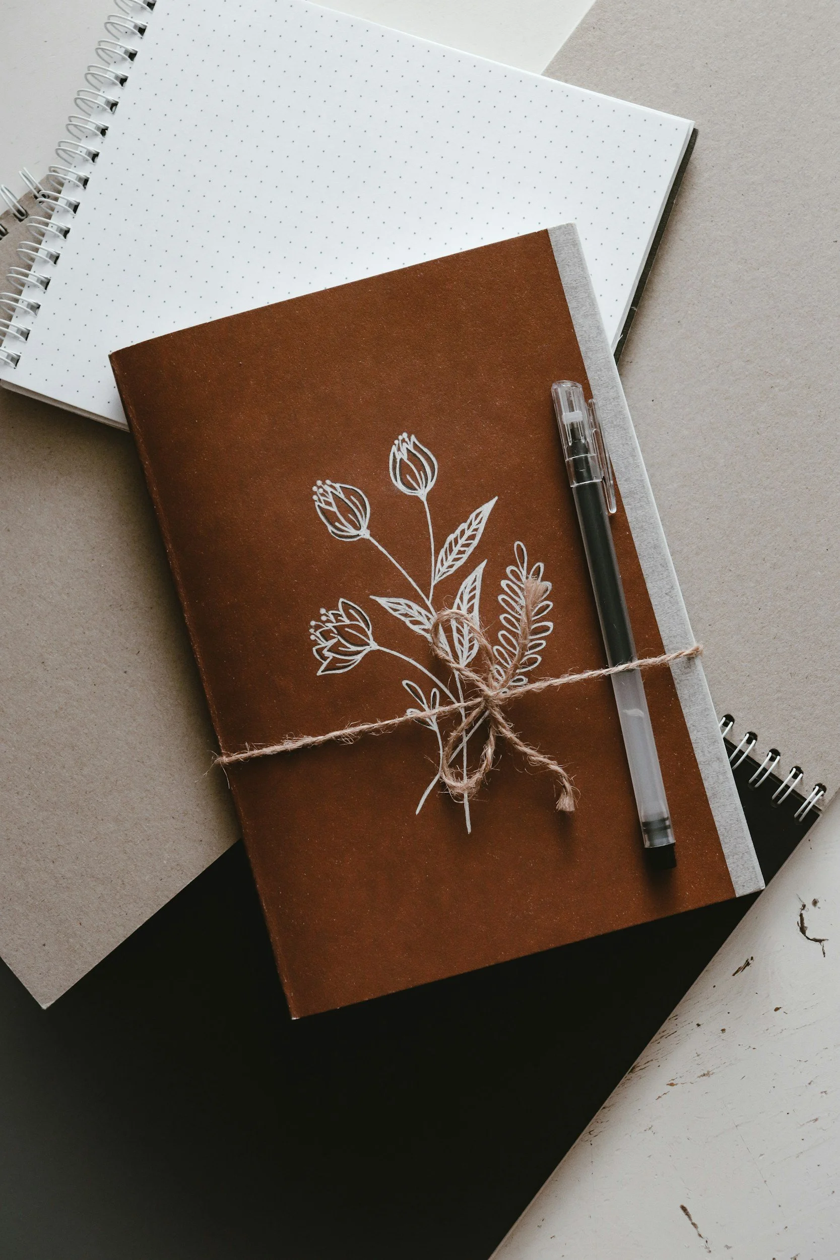 A brown notebook with flowers on the front and a pen tied with a string.