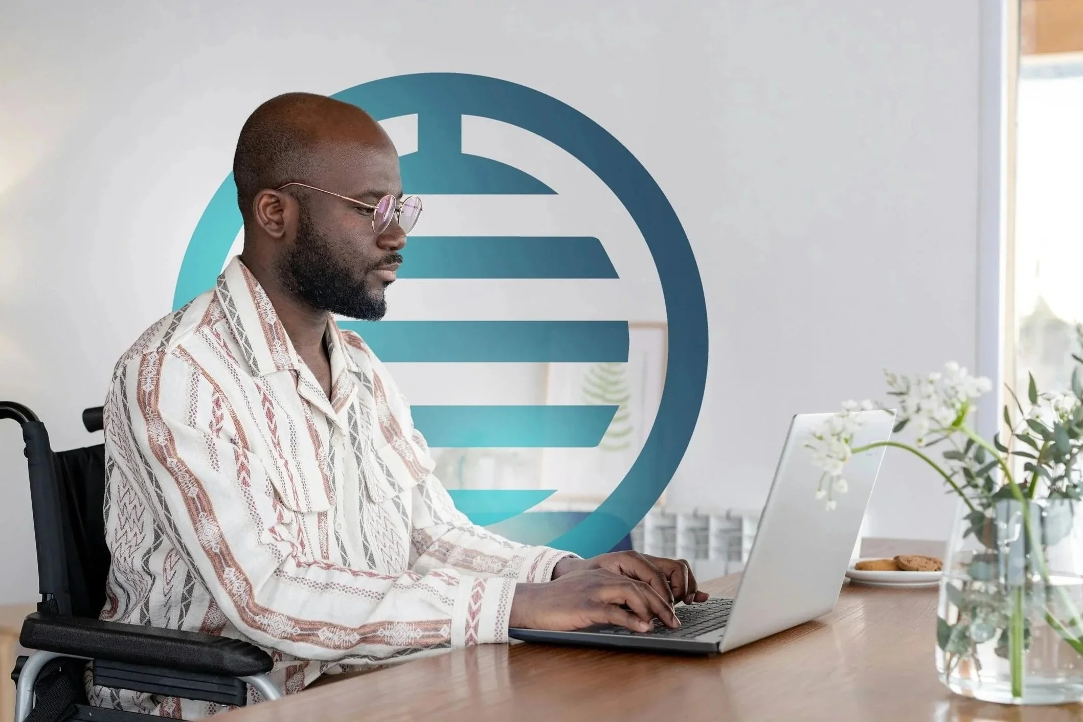 A man with glasses sitting in a wheelchair working on a laptop at a wooden table, with a vase of flowers and a plate of cookies nearby, in a bright room with a large logo in the background.