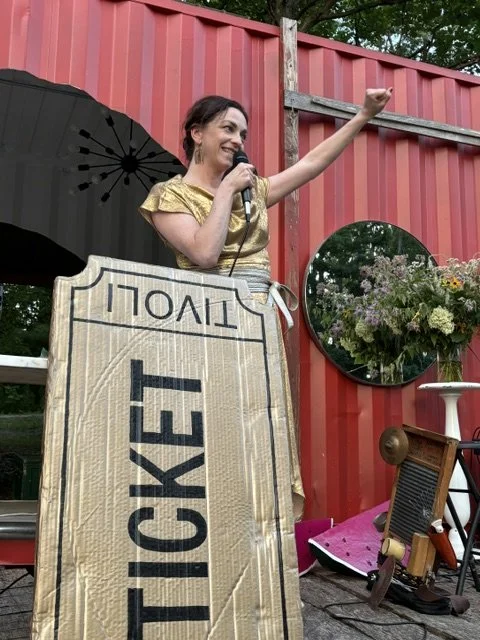 The Hudson Valley's best charity auctioneer and emcee, Karen Faith, in a gold dress speaking into a microphone at an outdoor event, standing behind a large gold ticket, with a red wall behind her.
