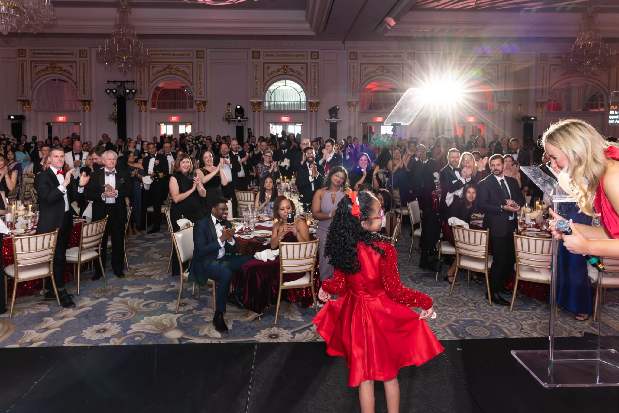 Crowd of people in formal attire clapping and smiling at an event in a decorated ballroom, with a young girl in a red dress at a podium encouraged by the worlds youngest charity auctioneer, Lyla Stebbins