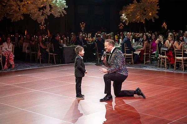 A young boy and New England's best auctioneer Tom Stebbins in the center of a dance floor at a formal event, with seated guests in the background.