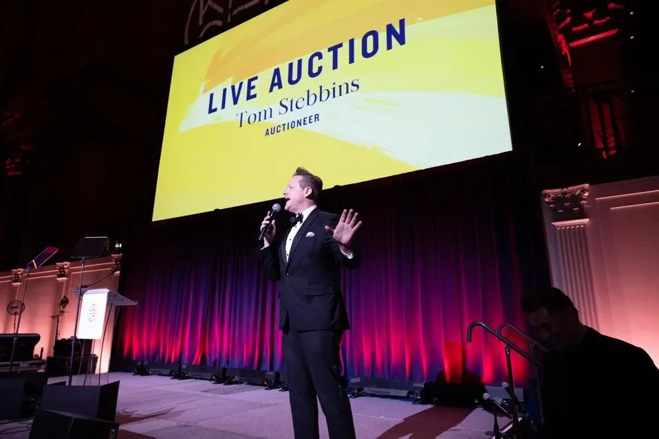Man in tuxedo speaking into microphone on stage during live auction, large screen behind him displaying 'LIVE AUCTION Tom Stebbins Auctioneer'.