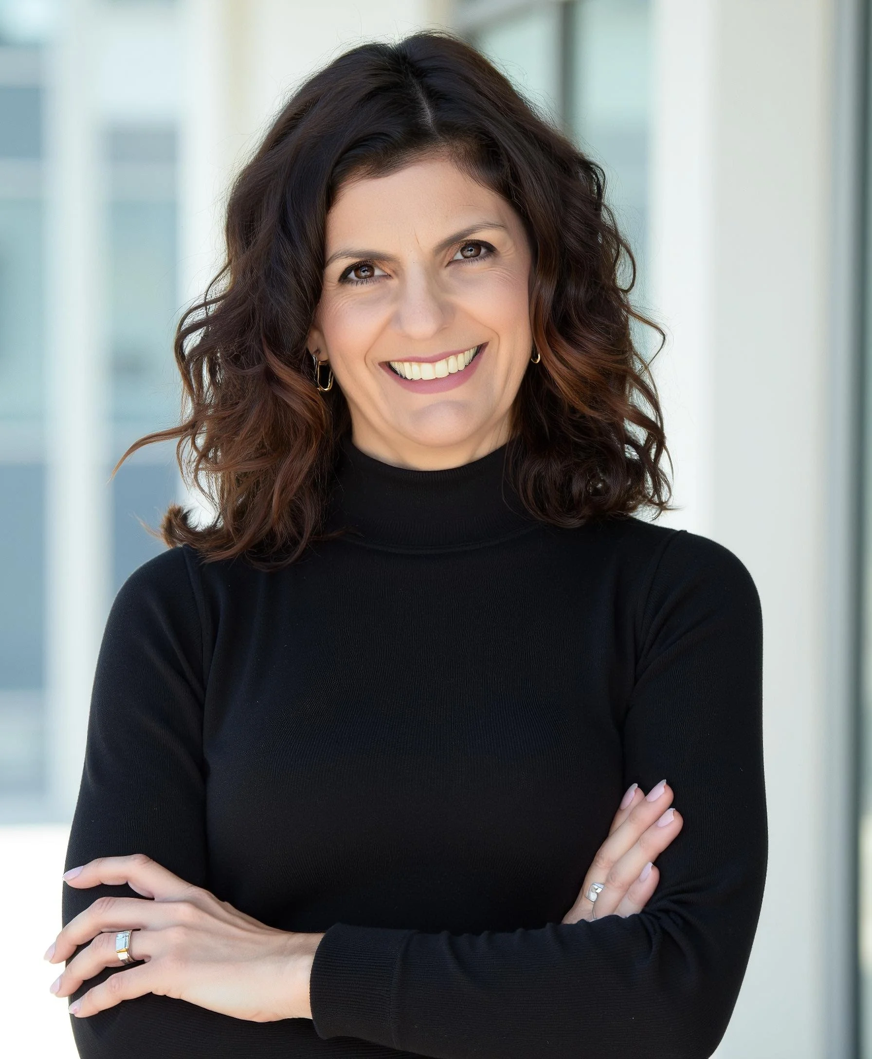 Dr. Mariela Shibley, a woman with dark, wavy hair wearing a black turtleneck sweater, smiling with crossed arms in front of a modern building window.
