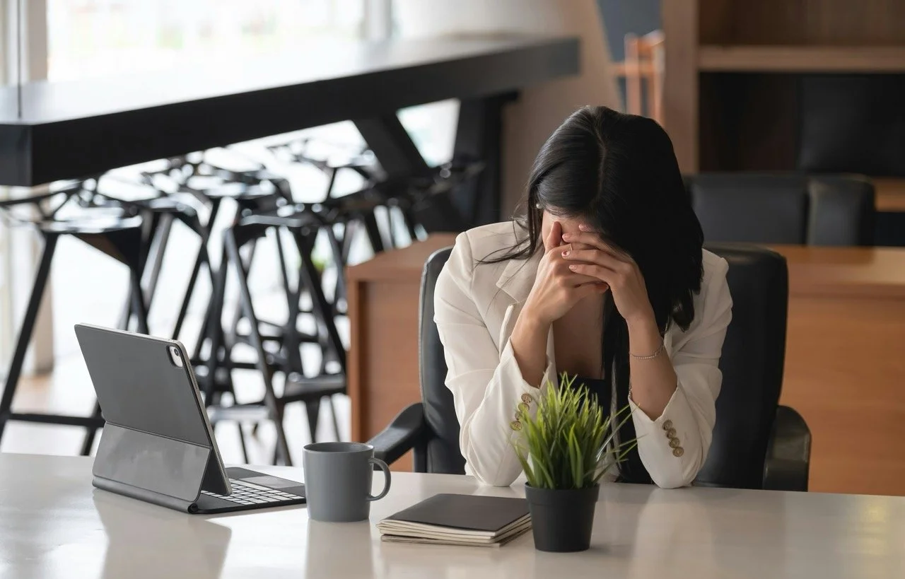 A woman in a white blazer sits at a desk with her head in her hands, appearing stressed or overwhelmed in a modern office setting. A tablet, coffee mug, notebook, and small plant sit on the desk beside her.