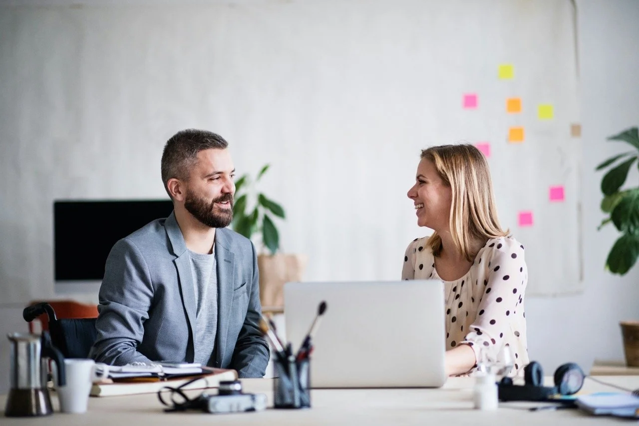 Two coworkers sit across from each other at a desk, smiling and talking during a collaborative meeting in a bright office.