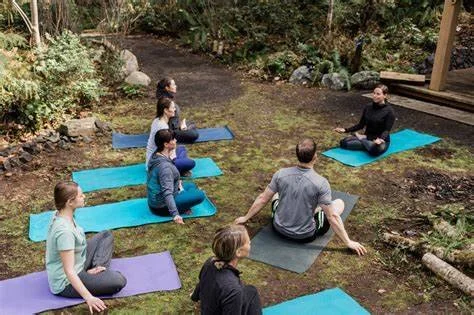 A group of people practicing yoga outdoors on a grassy area, sitting on yoga mats in front of a wooden building with trees in the background.