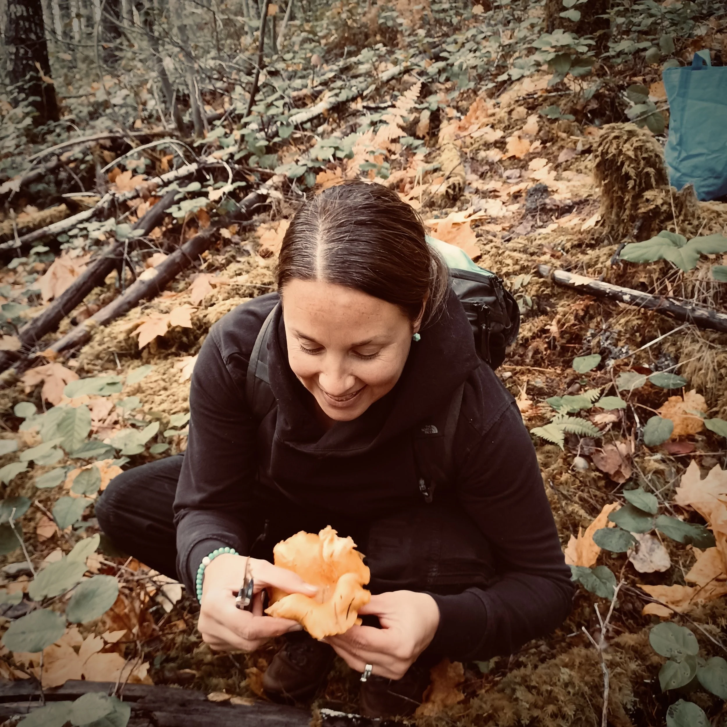 Michelle Pugh wearing a black knit beanie and black jacket smiling while holding a green leafy plant outdoors in a wooded area.