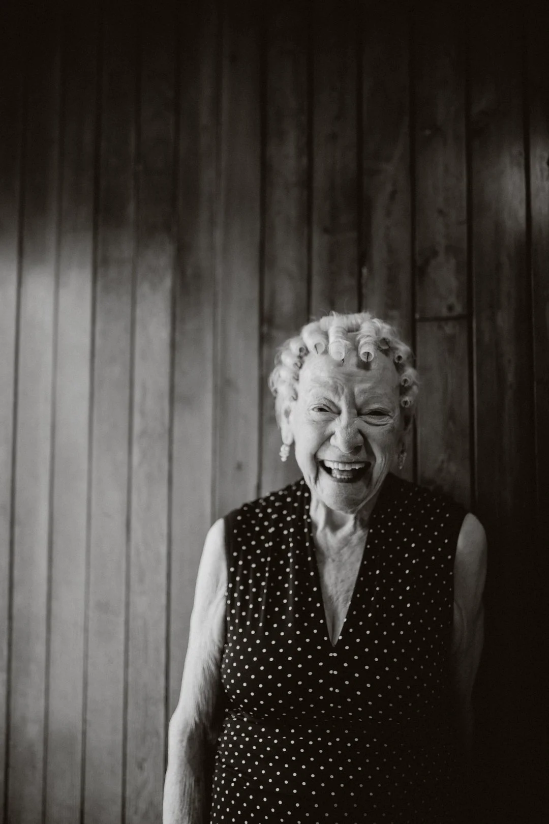 Elderly woman smiling with curlers in hair, wearing a polka dot dress, standing against wooden wall.