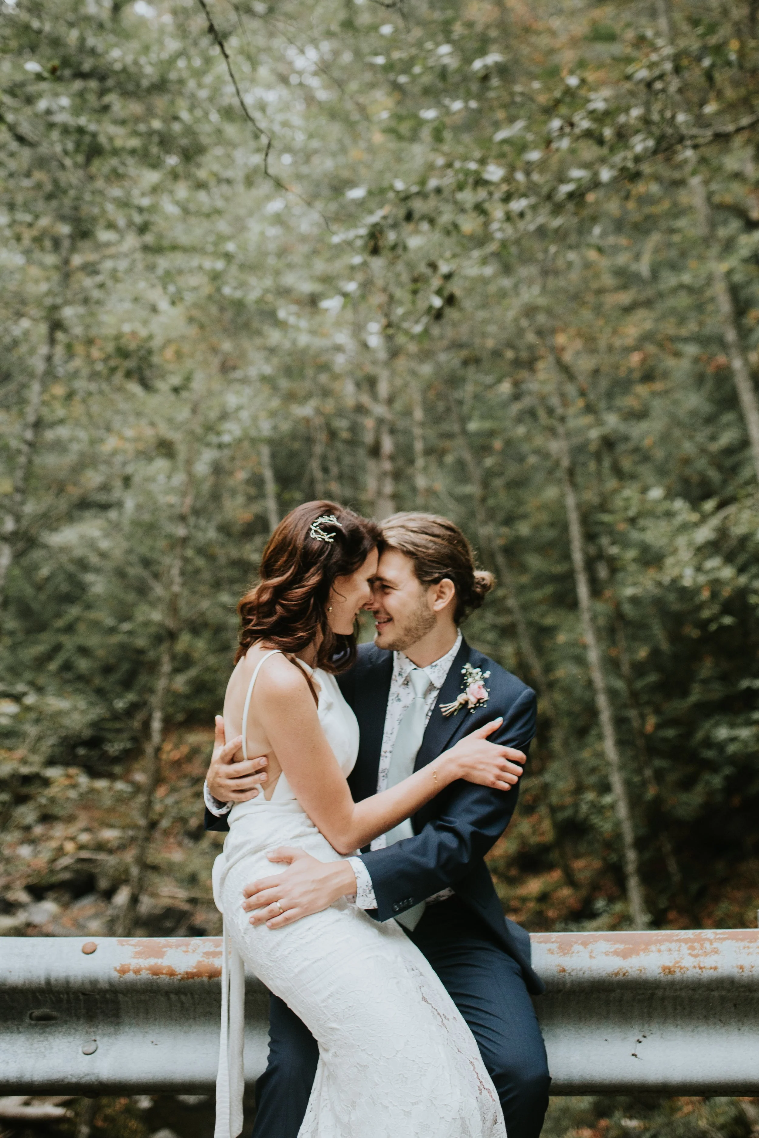 Bride and groom embracing in a forest setting, with the bride wearing a white dress and the groom in a navy suit, sitting on a guardrail.