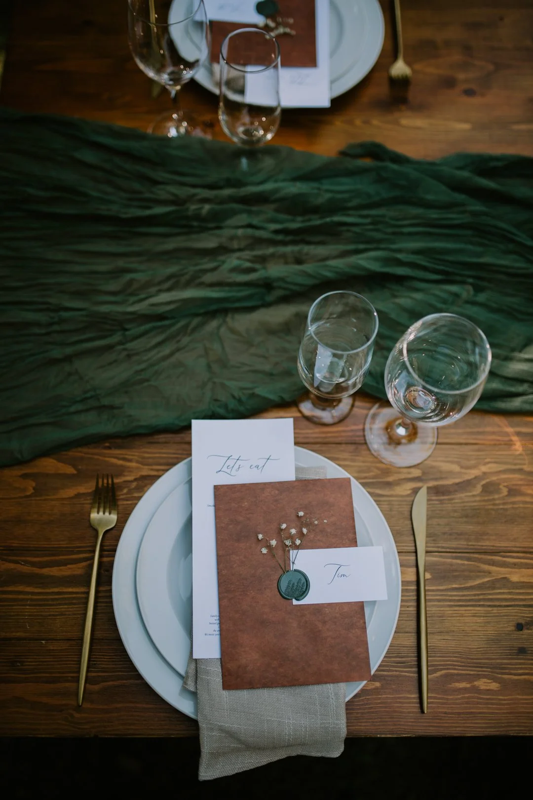 Elegant table setting with green tablecloth, white plates, gold cutlery, wine glasses, and a menu on brown and white paper with "Let's eat" written on it.
