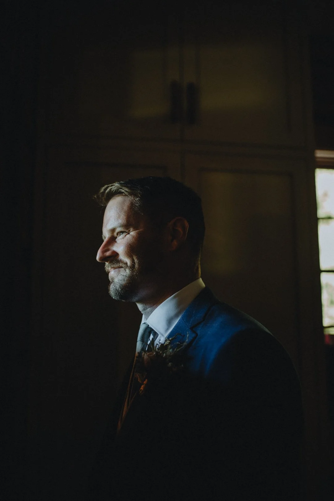 Man in a blue suit with a beard and mustache, illuminated by soft light, standing in a dimly lit room.