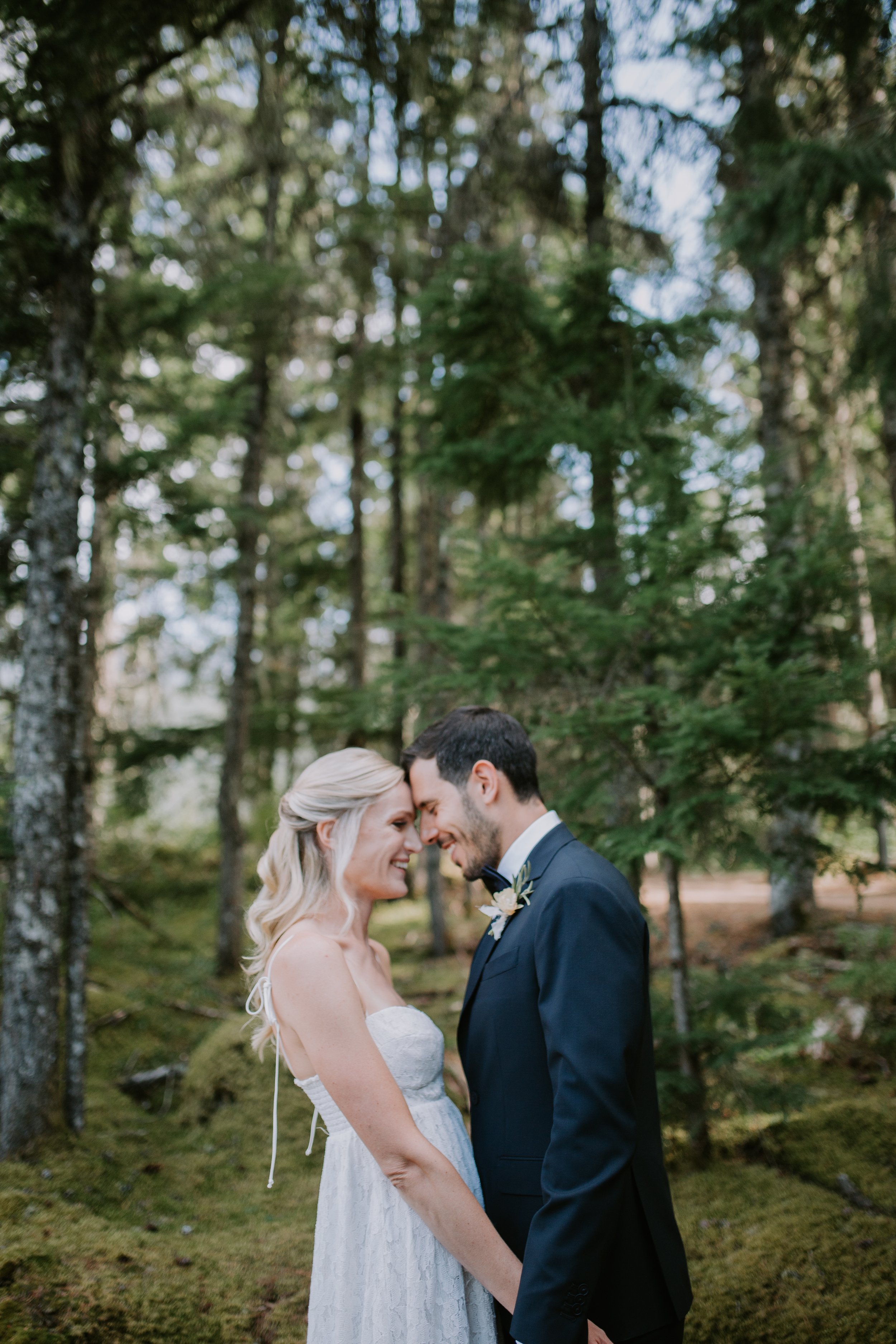 Bride and groom embracing in a forest setting, surrounded by green trees.