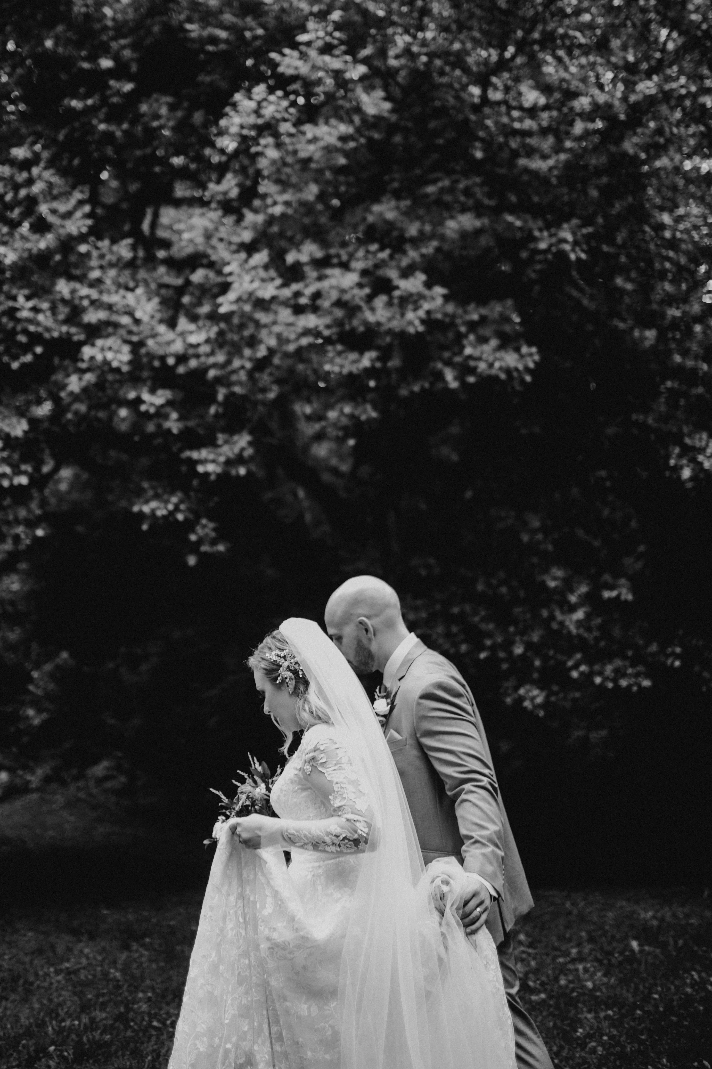 Bride and groom walking in a garden, bride in a lace dress and veil, black and white photo.