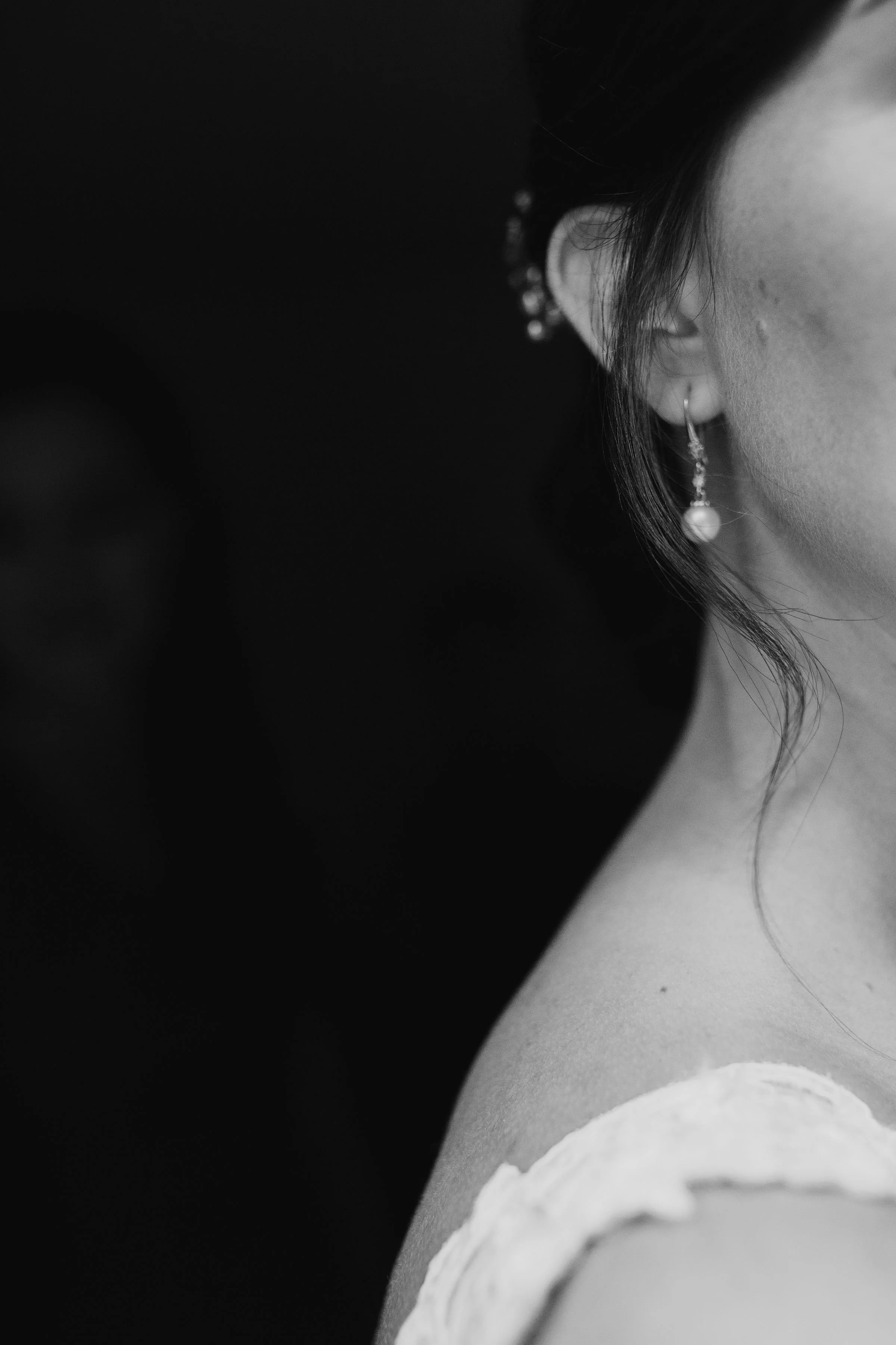Black and white close-up of a woman's neck and shoulder, showing an earring and lace dress detail.