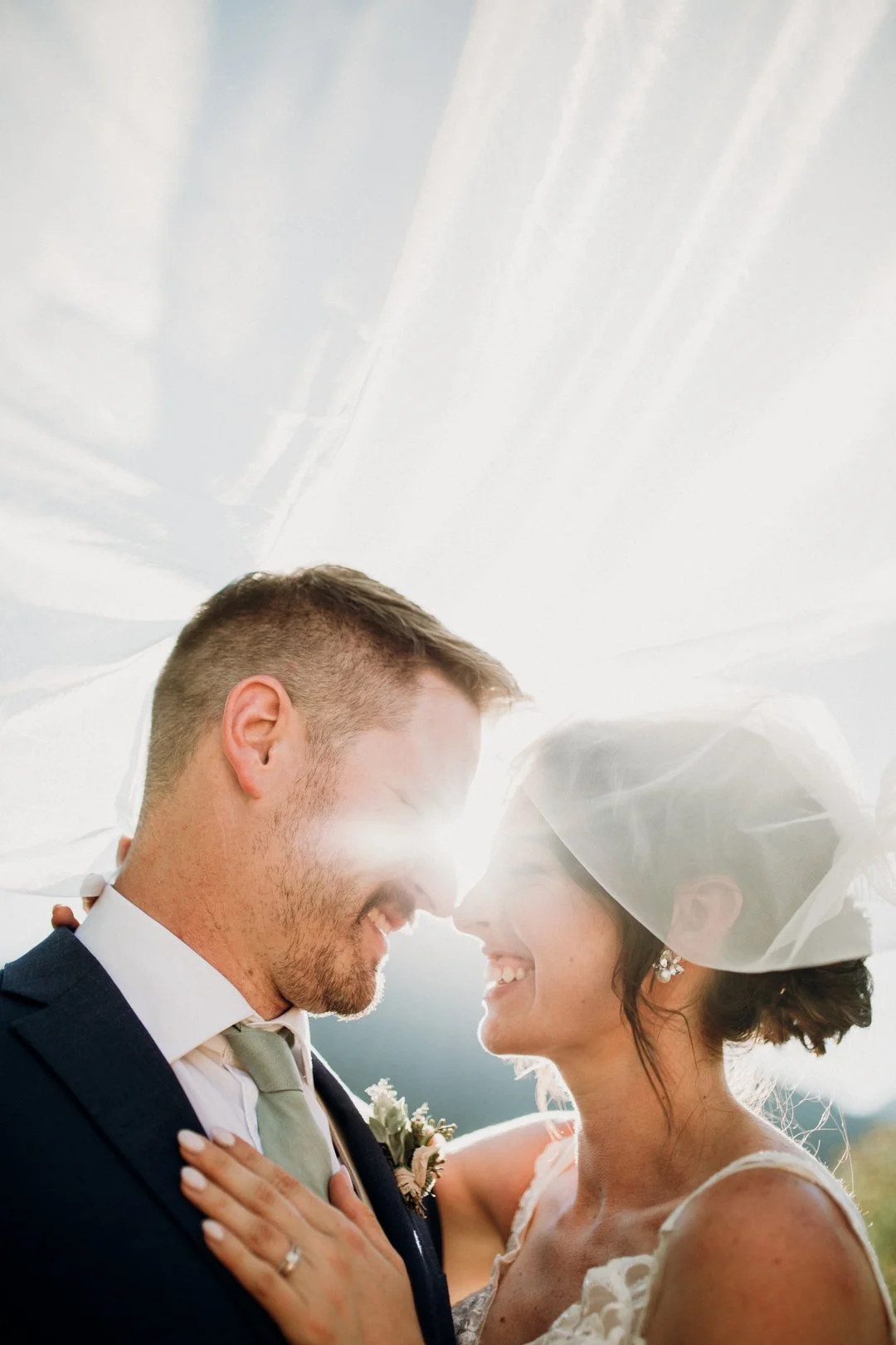 A bride and groom smiling at each other with sunlight behind them. The bride is wearing a veil and the groom is in a suit.