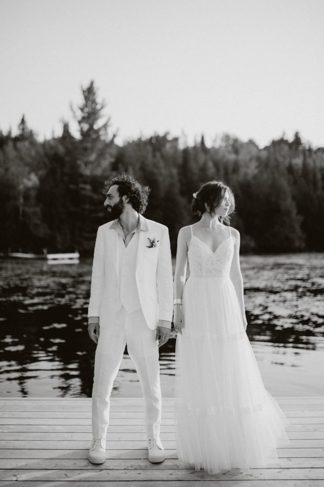 A black and white photo of a bride and groom standing on a wooden dock by a lake, holding hands, with a forest in the background.