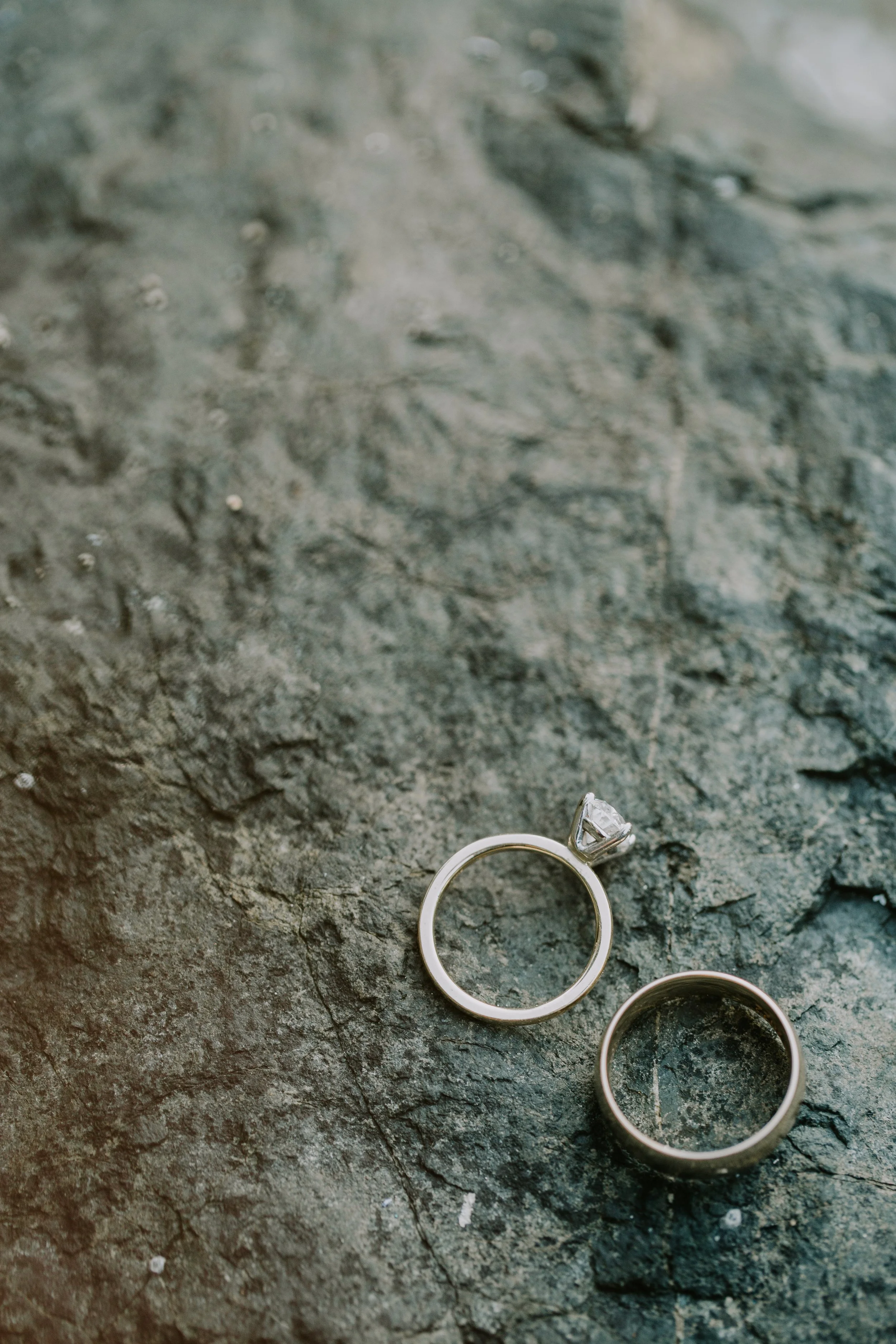 Two rings, an engagement ring with a diamond and a plain wedding band, placed on a textured stone surface.