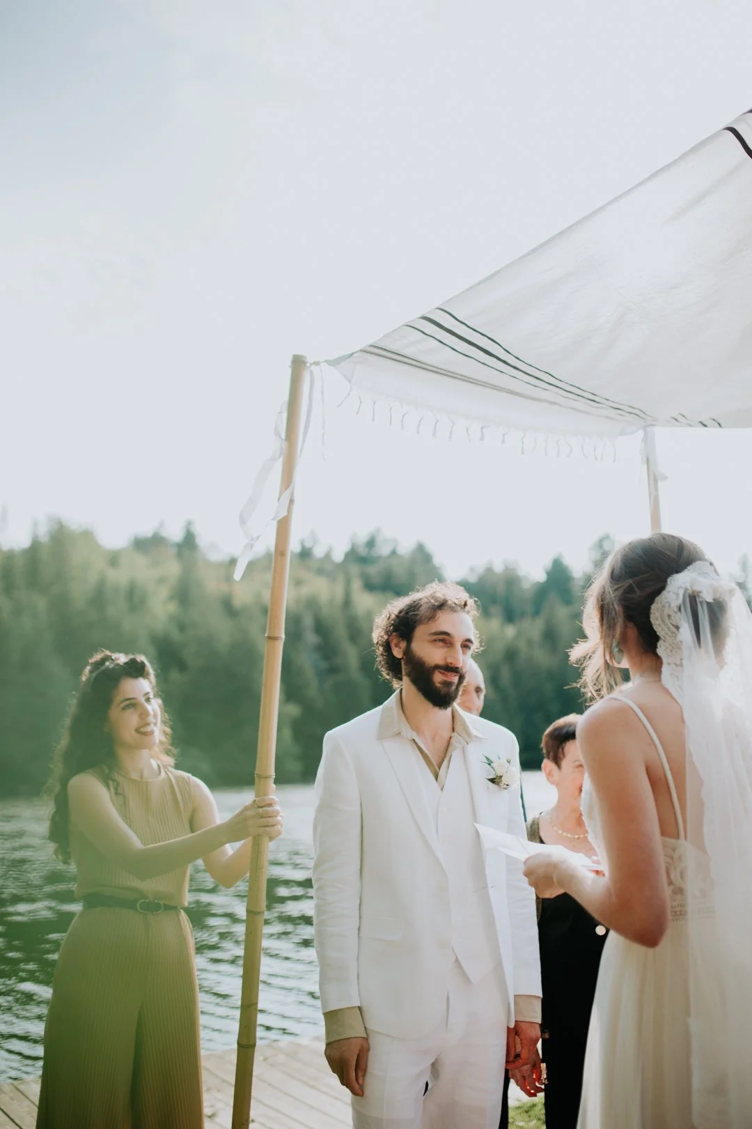 A couple getting married outdoors under a chuppah near a lake with trees in the background. The groom is wearing a white suit and the bride is in a white dress with a veil. A person is holding one of the chuppah poles.