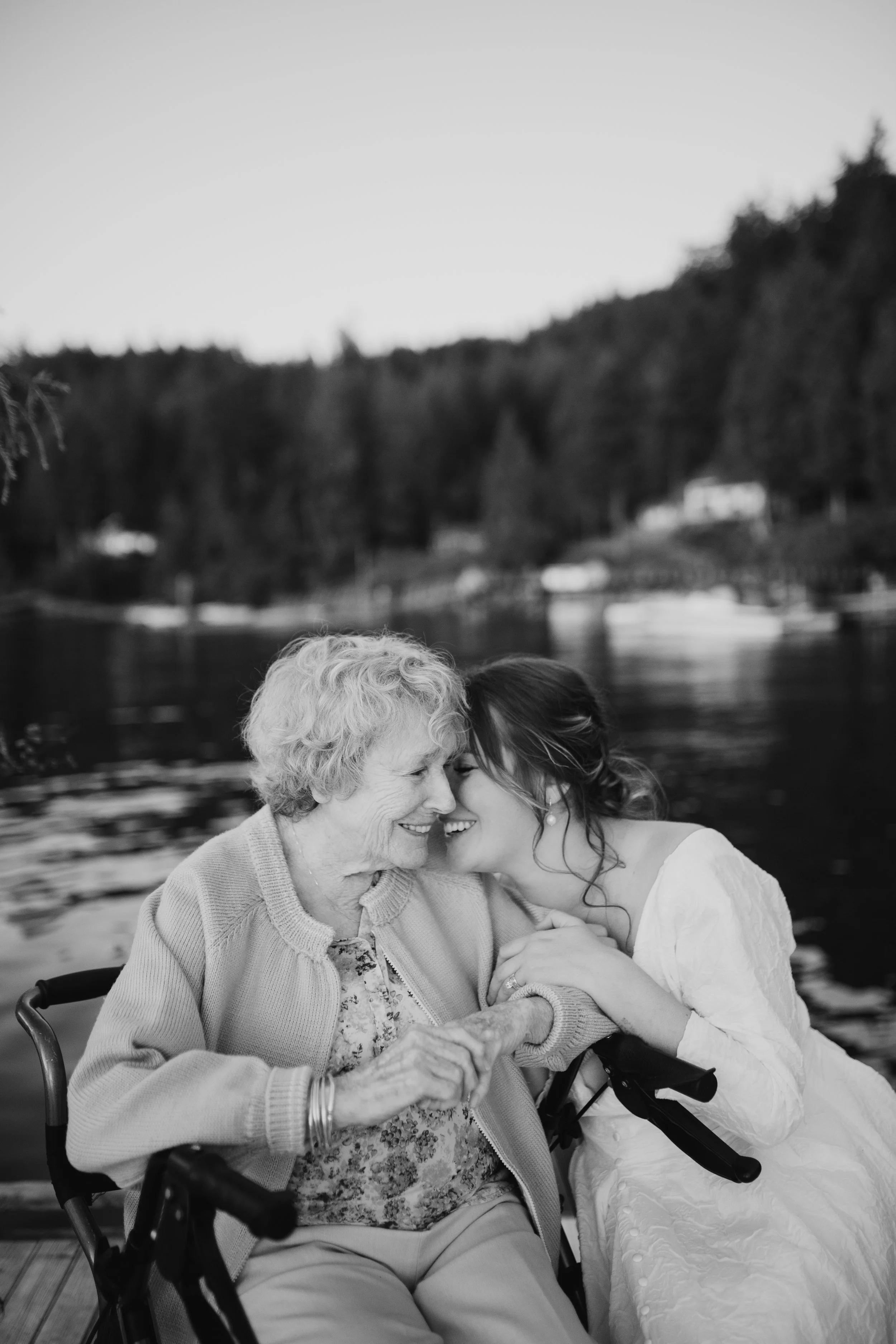 Elderly woman and young woman smiling and touching foreheads, beside a scenic lake, in black and white.