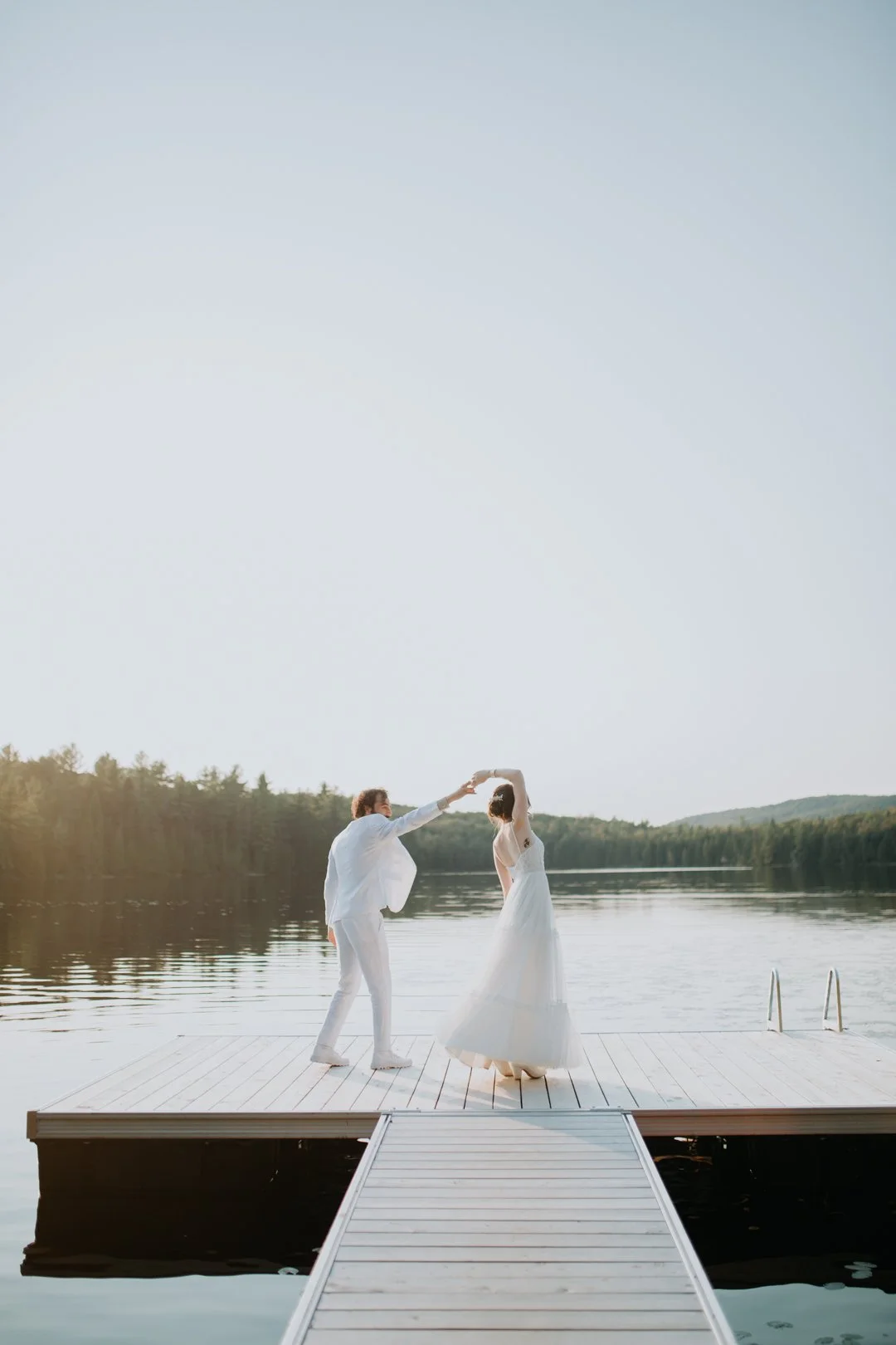 A couple in wedding attire dancing on a wooden dock by a lake with a forest in the background.