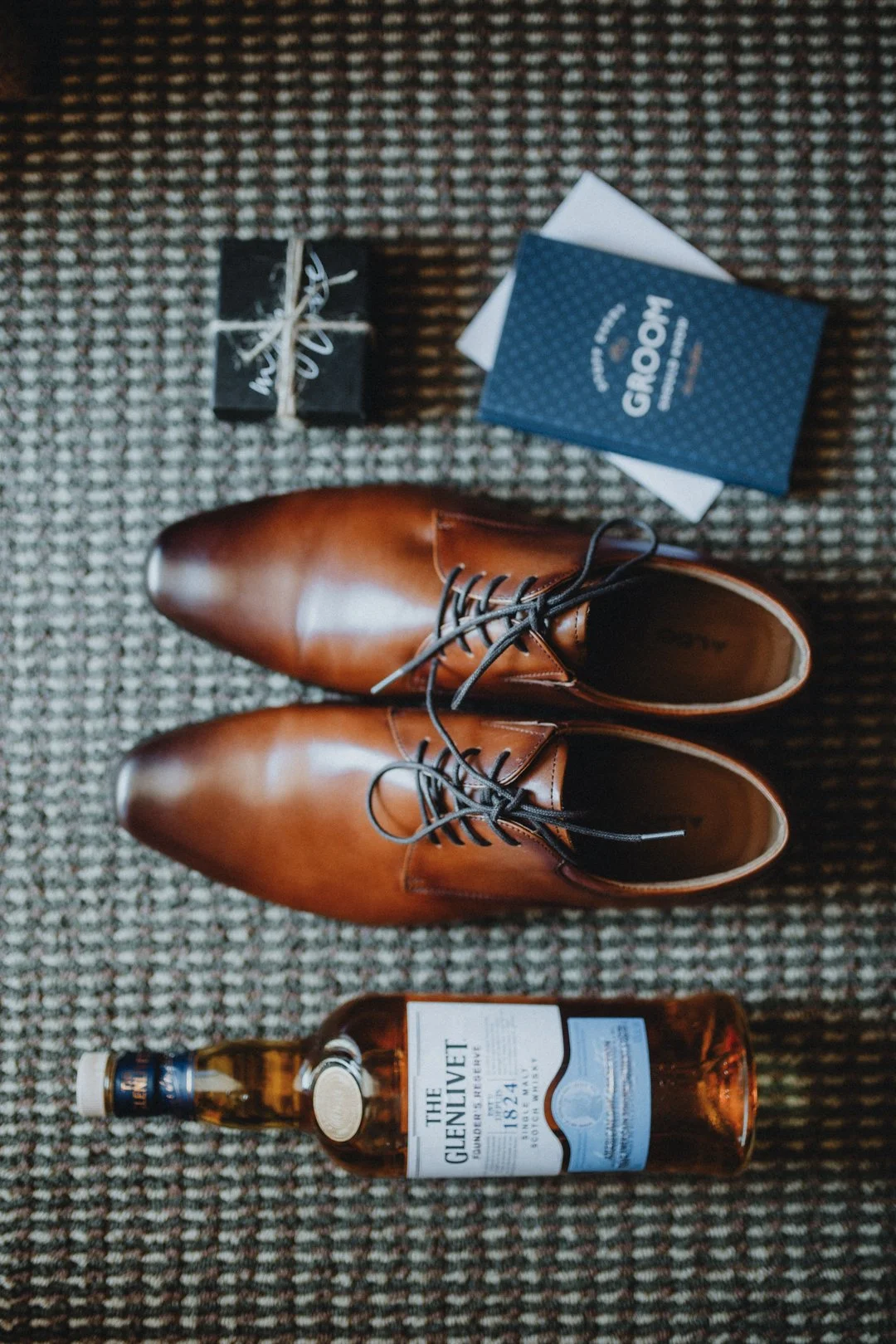 Brown leather dress shoes, a bottle of The Glenlivet whisky, a small black gift box tied with string, and a groom card on a patterned carpet.