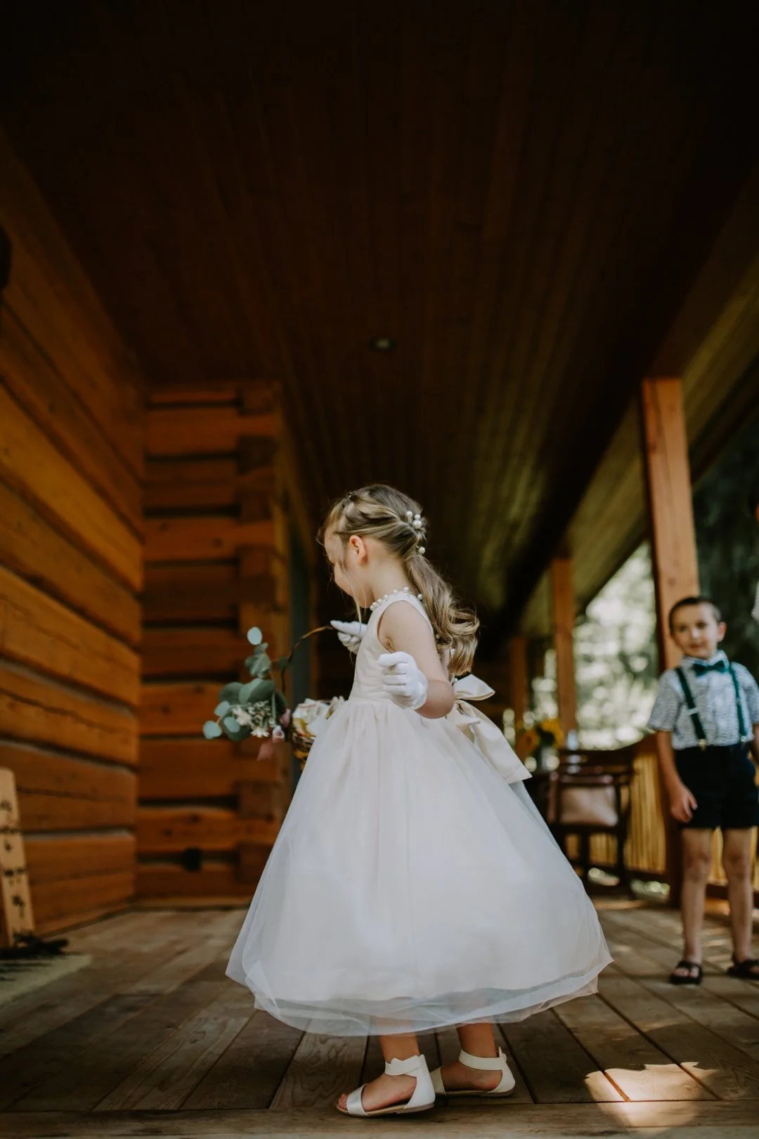 Young girl in white dress on wooden porch, holding flowers, with boy in background wearing suspenders.