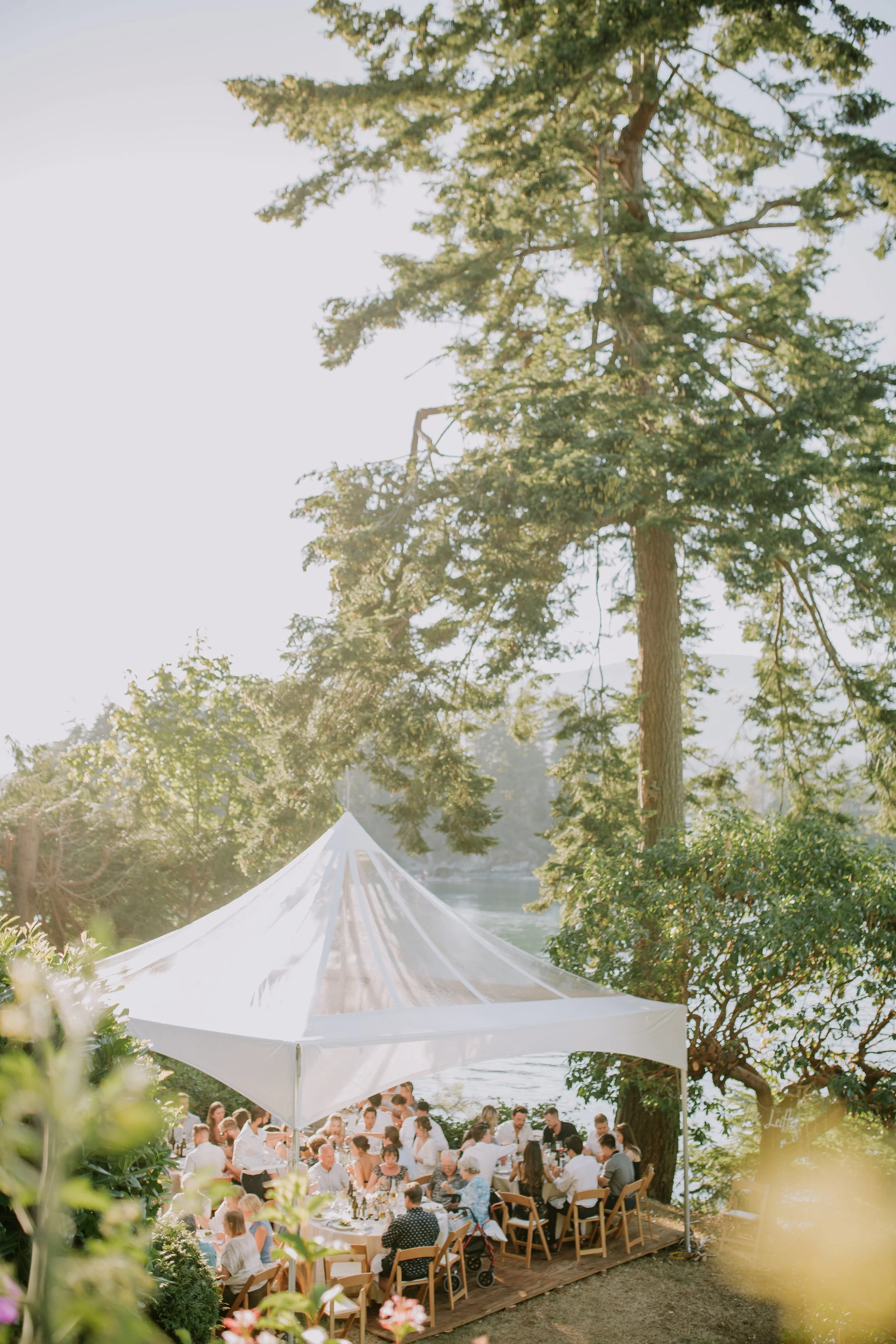 Outdoor gathering under a white tent next to a lake, surrounded by tall trees, with people seated at tables during a sunny day.