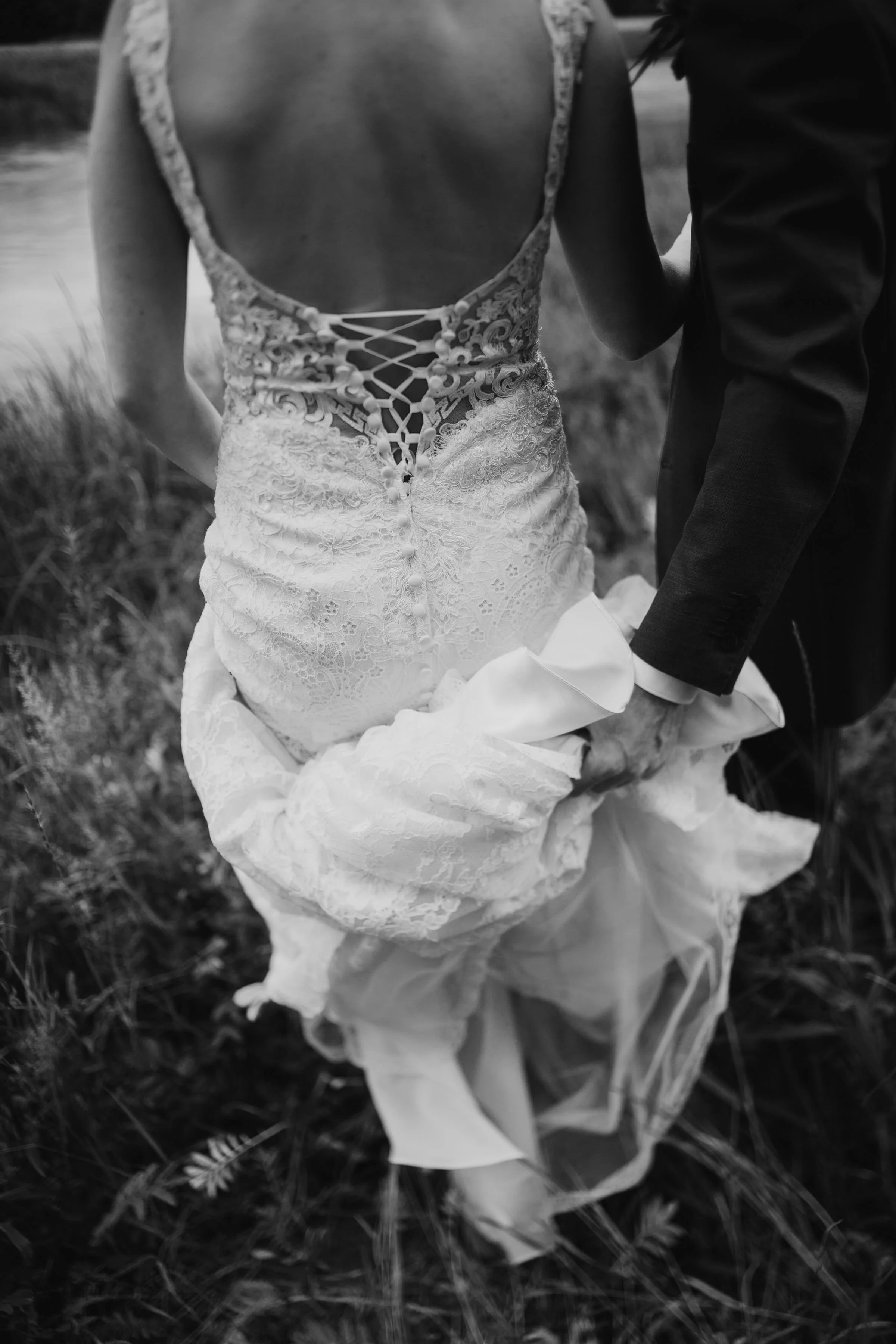 Bride with lace-back wedding dress holding gown, accompanied by someone in a suit, walking outdoors in grassy area.