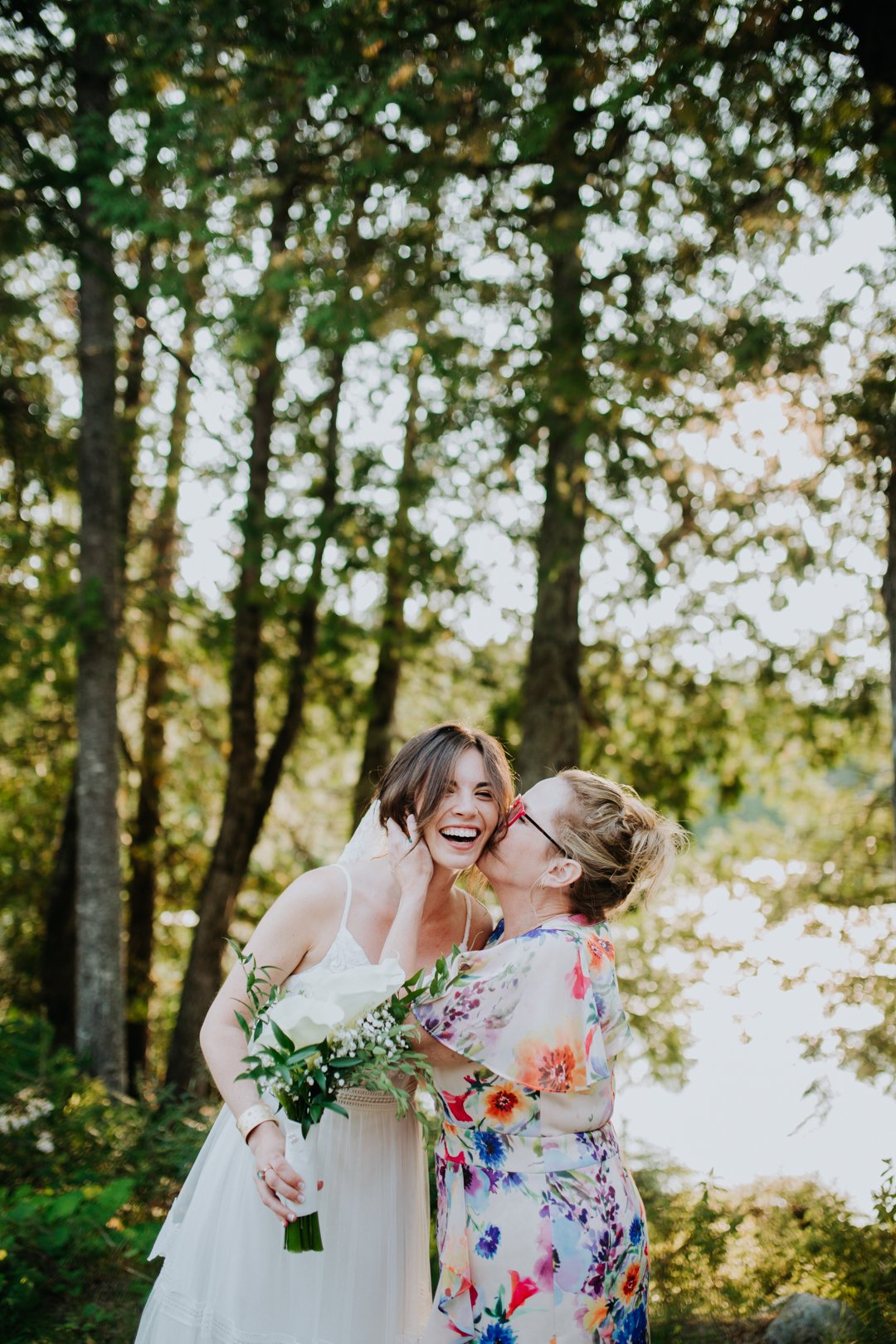 Two women laughing outdoors, one in a white dress holding a bouquet, and the other in a colorful floral outfit, surrounded by trees.