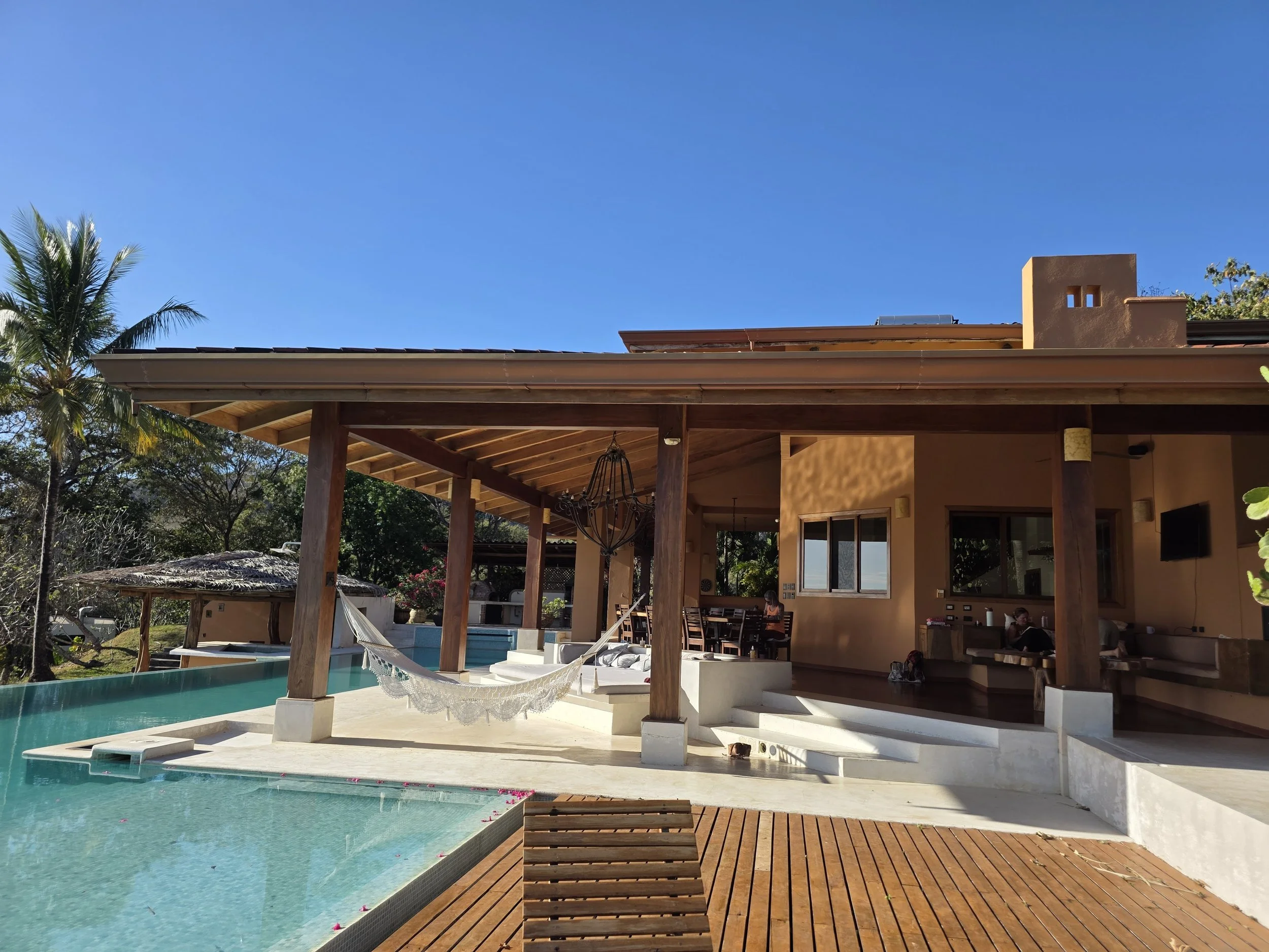 A backyard scene with a pool, hammocks, a covered patio area, and a house with a beige stucco exterior and a chimney, surrounded by trees and clear blue sky.
