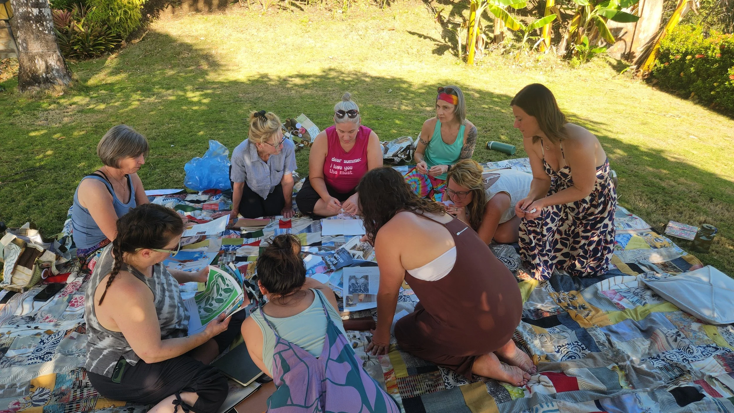 A group at the women's ayahuasca retreat sitting together outdoors on a blanket in a sunny garden, engaging in arts and crafts activities with paper, magazines, and other materials.