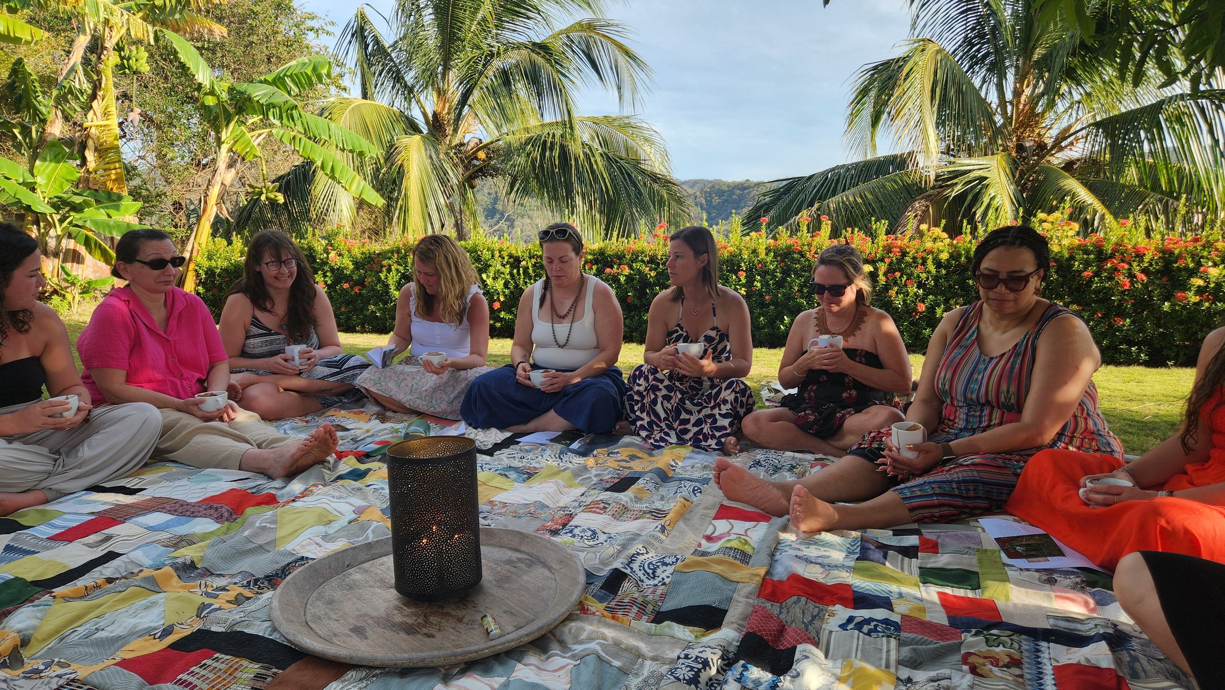 A group of women sitting on a blanket outdoors surrounded by lush tropical plants and trees, holding cups, possibly enjoying a tea or coffee gathering in a garden.